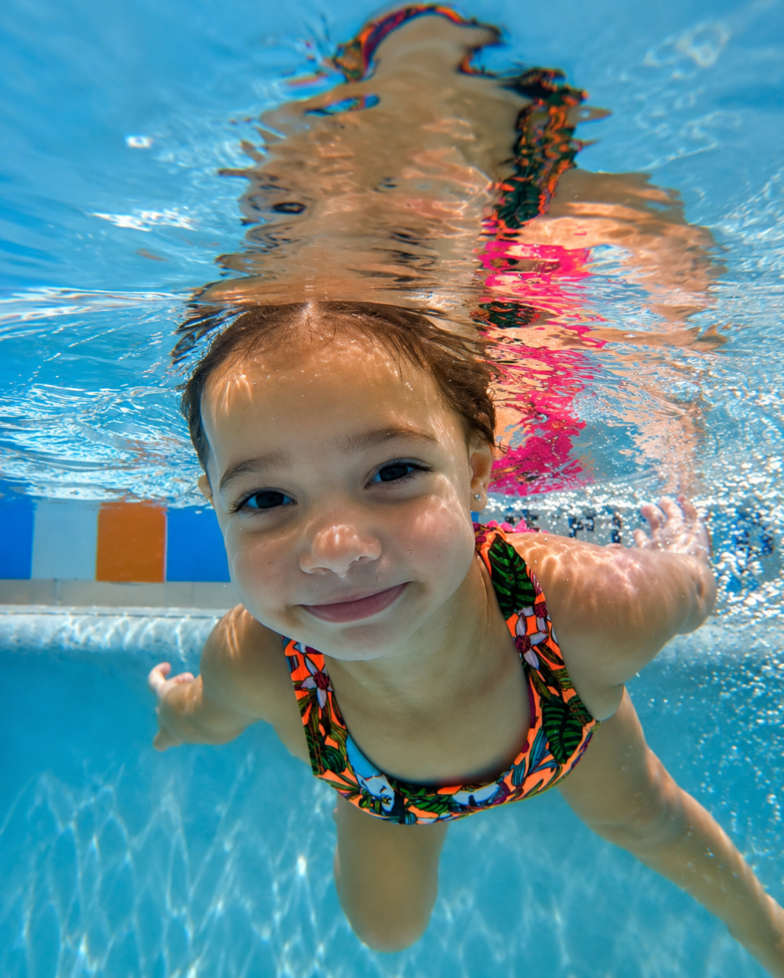 Young girl with a big smile swimming underwater in a pool, wearing a colorful swimsuit.