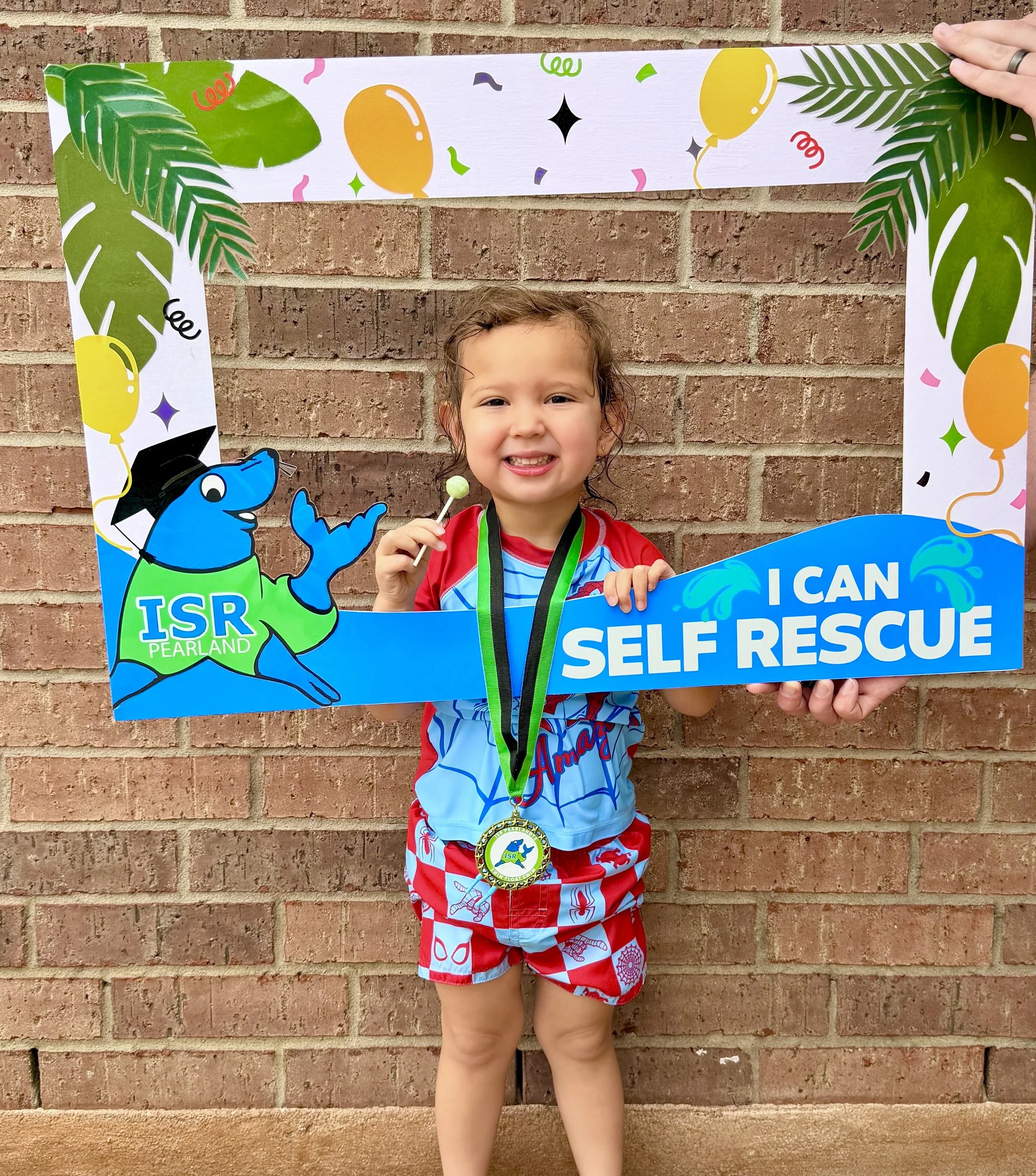 Young child standing in front of a brick wall, holding a green lollipop, wearing a red and blue swimsuit with cartoon lobster and crab patterns, and a medals around their neck. There is a colorful, celebratory border frame with balloons, leaves, and confetti.