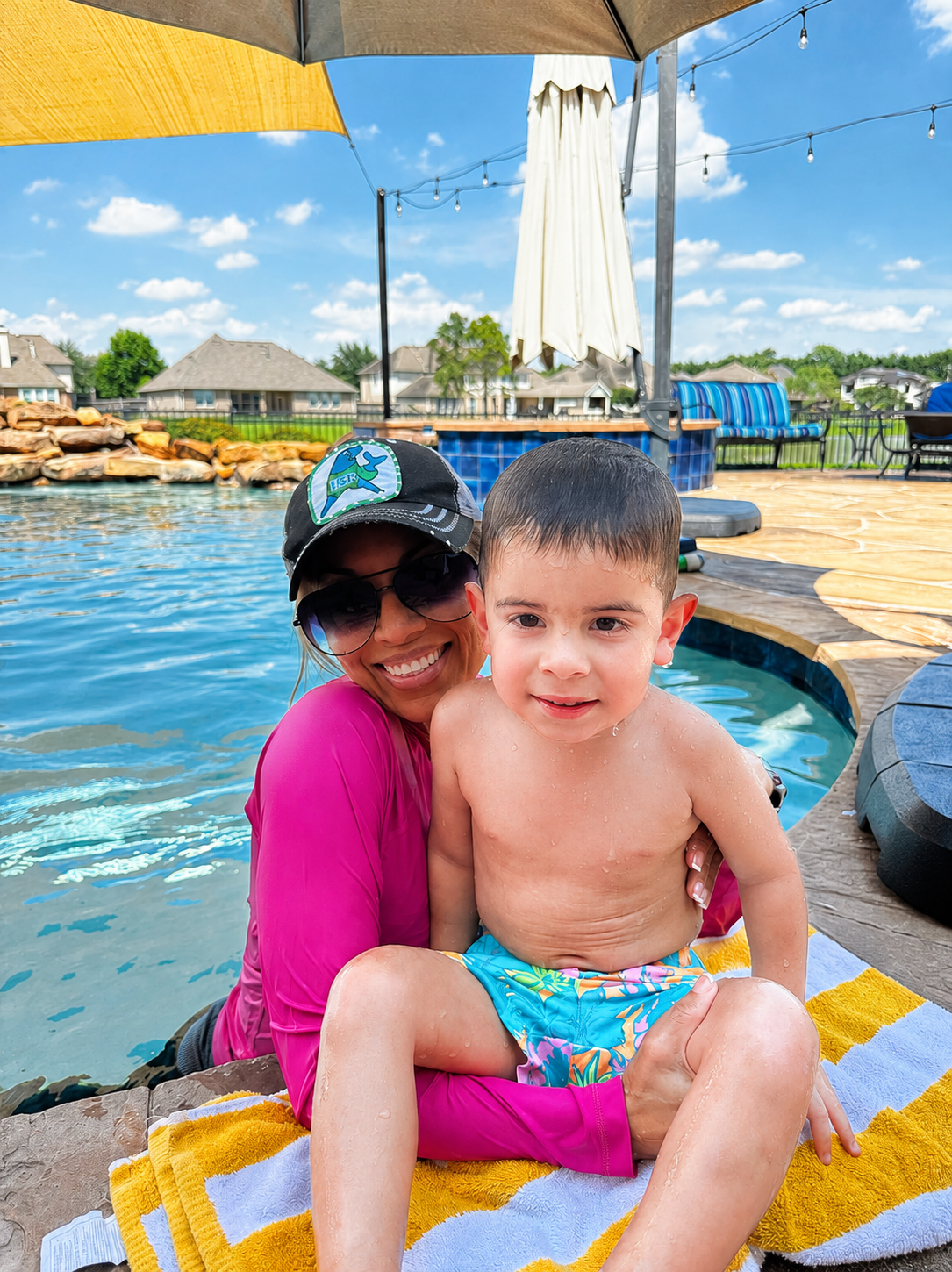 A woman and a young boy sitting by a swimming pool, smiling at the camera. The woman is wearing sunglasses, a hat, and a pink shirt, while the boy is shirtless and wearing colorful swim trunks. Behind them is a pool with a rocky waterfall, umbrellas, and outdoor furniture on a sunny day.
