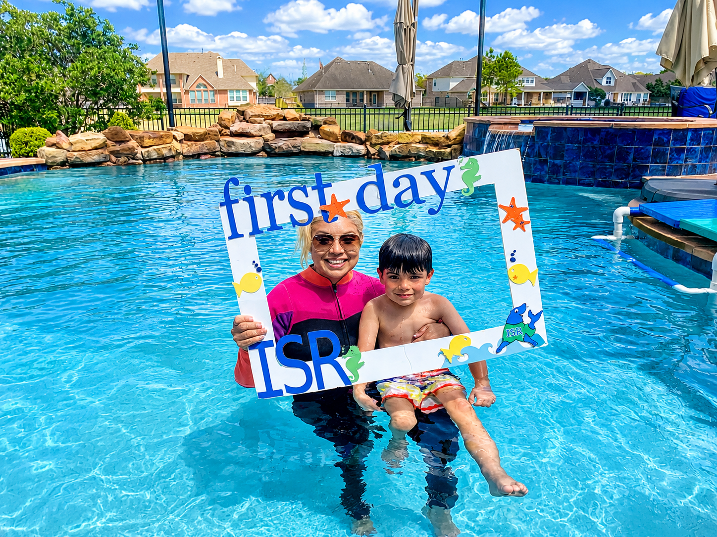 Woman and boy in a swimming pool holding a sign that says 'first day' and 'ISR' with fish and seahorse decorations, on a sunny day with clear blue sky and neighborhood houses in the background.