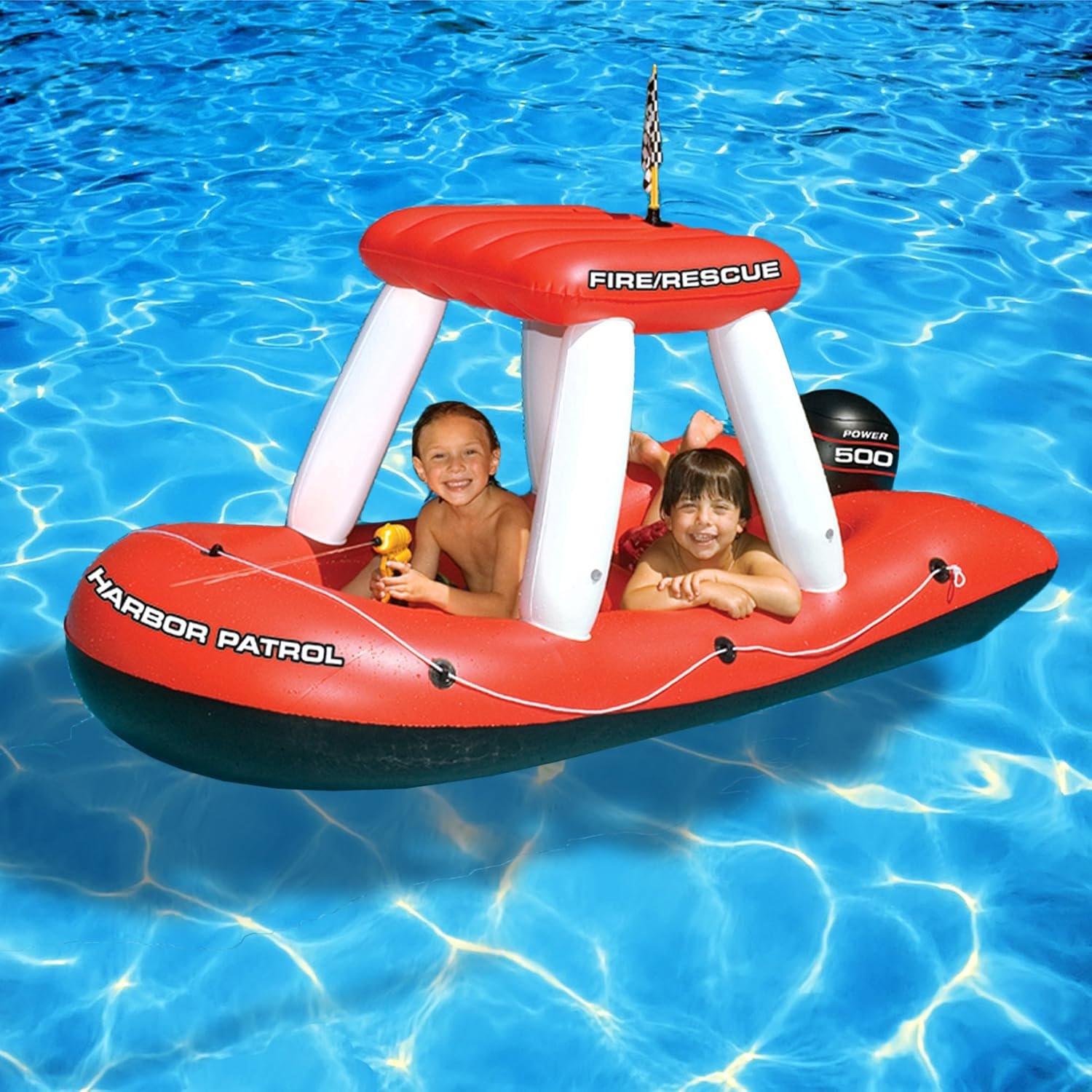 Two children are happily riding in a red and white inflatable boat labeled 'Harbor Patrol' on blue water. One child is holding a small yellow toy, and the boat features a small black and yellow checkered flag on top.