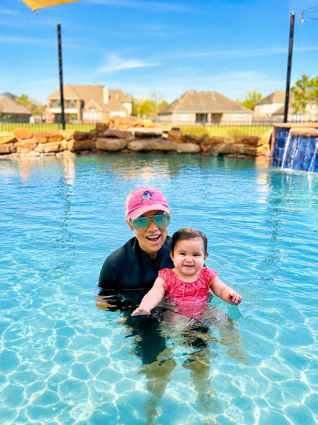 A woman and a young girl are smiling and enjoying themselves in a backyard swimming pool. The woman is wearing sunglasses, a pink cap, and a dark swim shirt, while the girl wears a pink swimsuit and has wet hair.