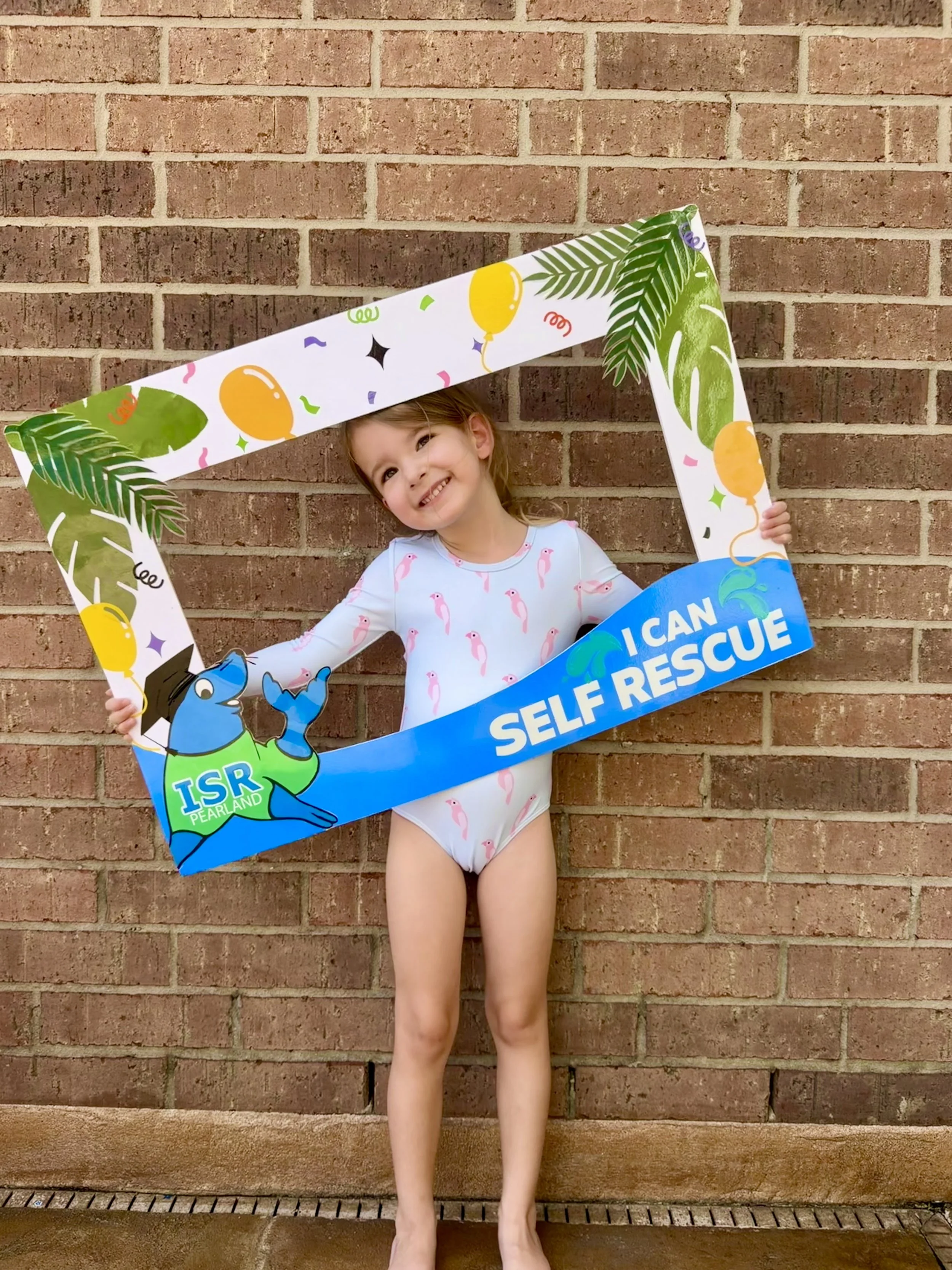 A young girl standing in front of a brick wall, holding a colorful photo frame with the words 'I CAN RESCUE' and illustrations of a blue dog wearing a firefighter hat and a yellow balloon, celebrating self-rescue.
