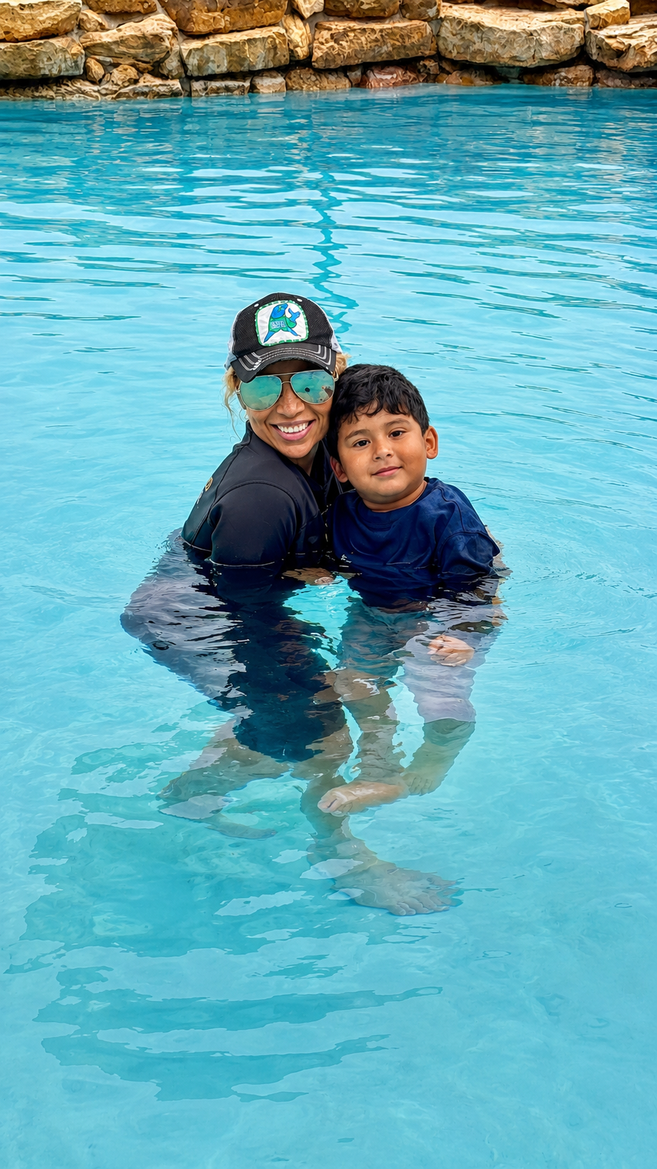 A woman and a young boy are standing in a swimming pool, smiling and hugging. The woman is wearing sunglasses and a cap, while the boy is in a blue shirt. There are rocks along the pool's edge in the background.