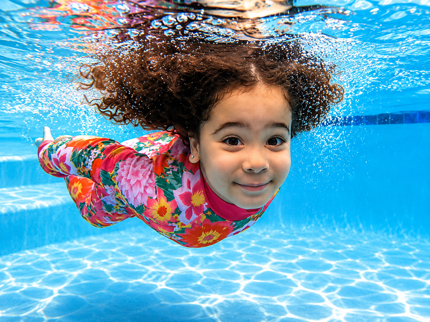 A young girl swimming underwater in a swimming pool, smiling and wearing a colorful floral rash guard.