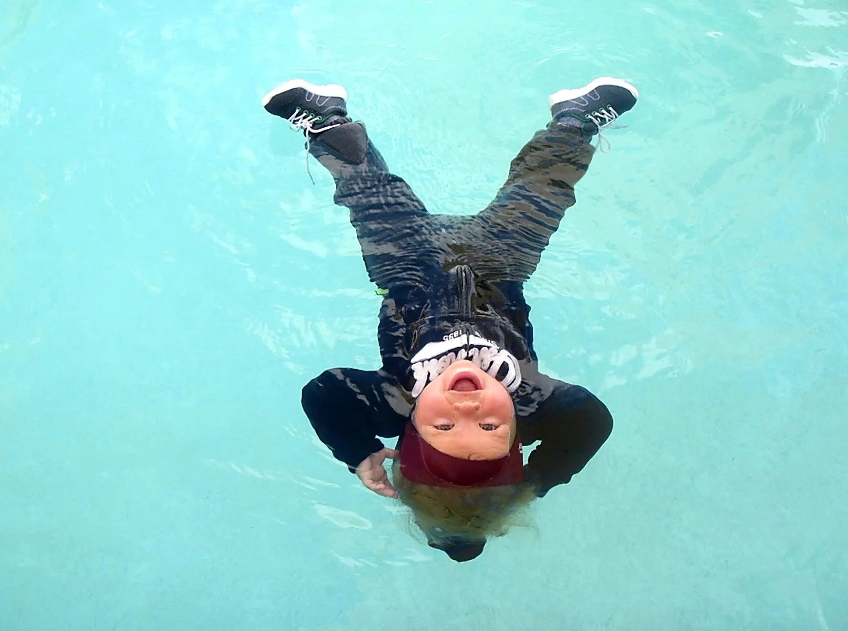 Child lying on back in a swimming pool, smiling and wearing a black swimsuit, a red swimming cap, and a black life vest.