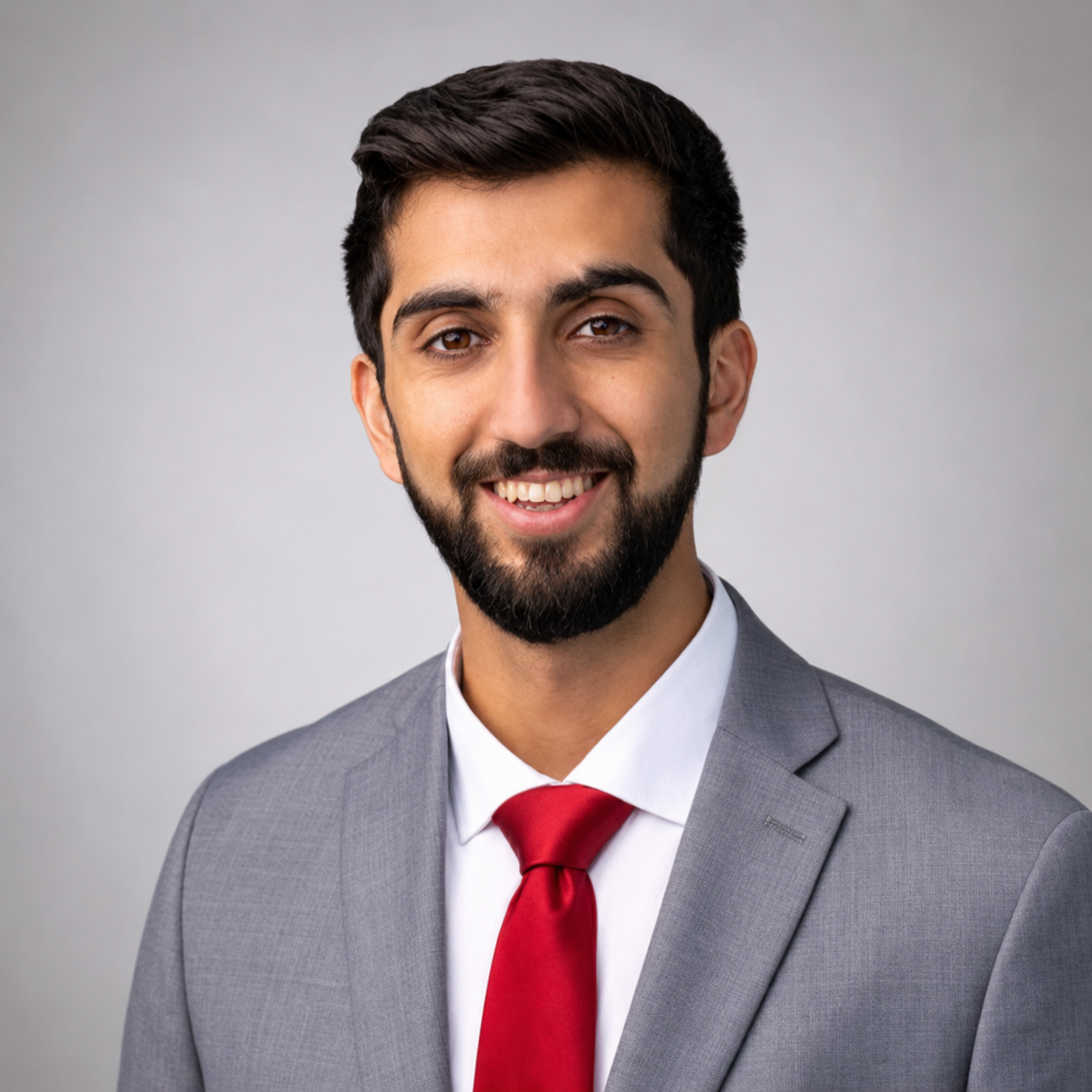 Professional headshot of a man in a gray suit, white shirt, and red tie, smiling with a beard and short dark hair, against a gray background.