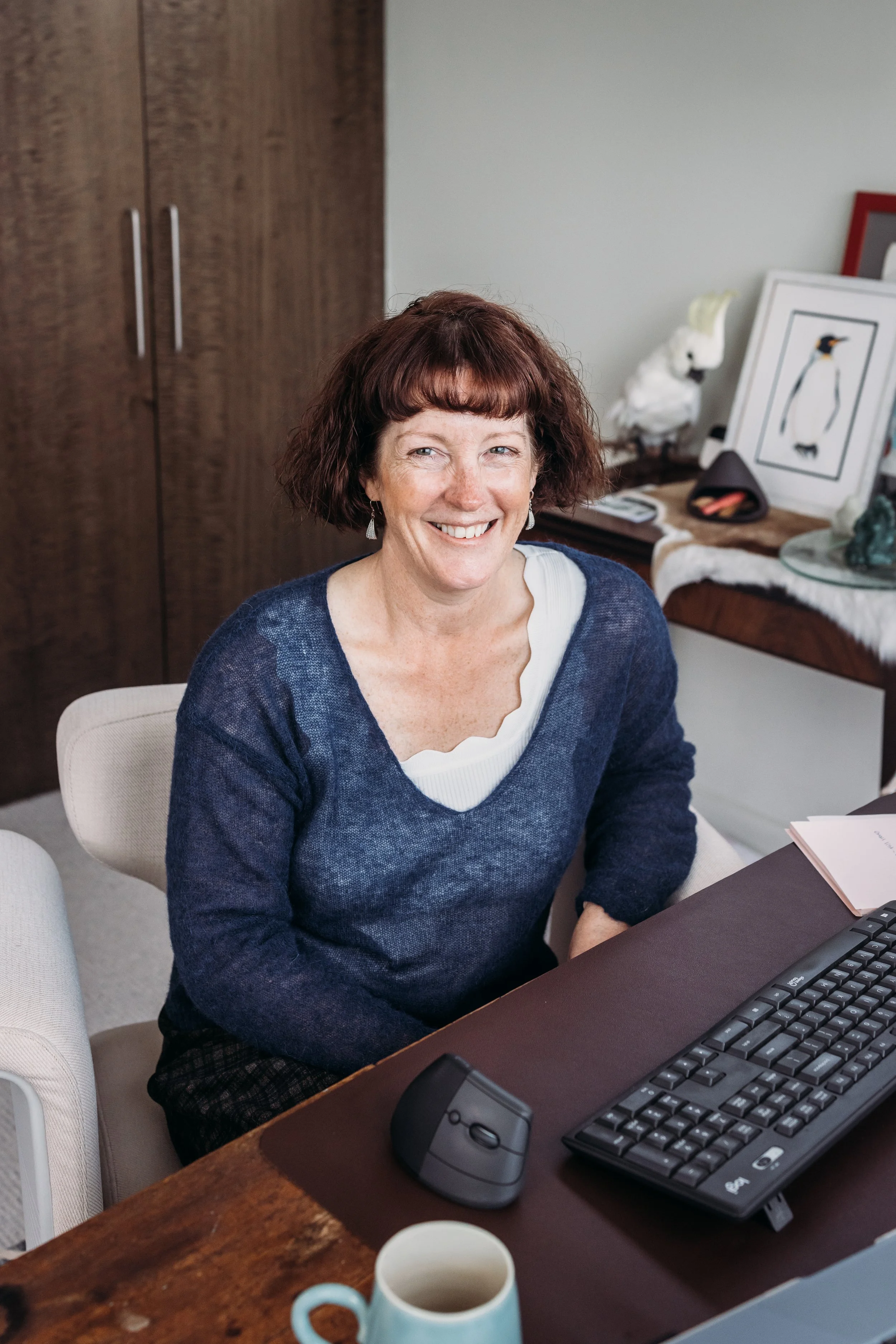 A smiling woman with shoulder-length brown hair and bangs, wearing a navy sweater and earrings, sitting at a desk with a computer keyboard and mouse, in a room with framed pictures and a bird sculpture in the background.
