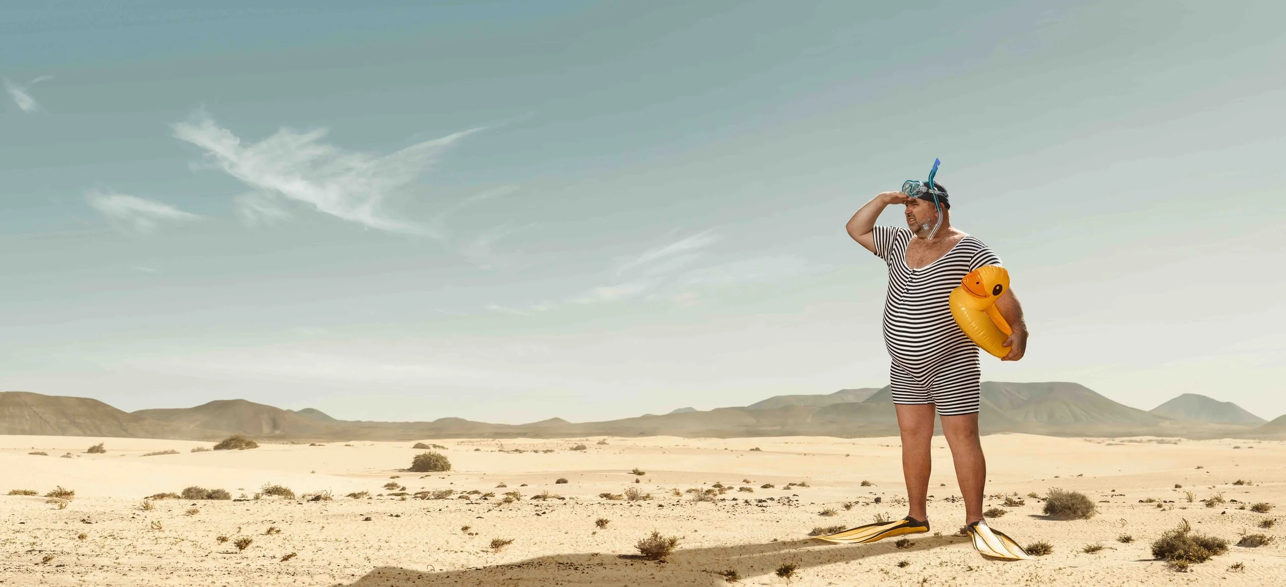A man in striped clothing with snorkeling gear, a yellow inflatable ring, and flippers stands on a sandy desert landscape with mountains in the background, saluting as he looks into the distance.