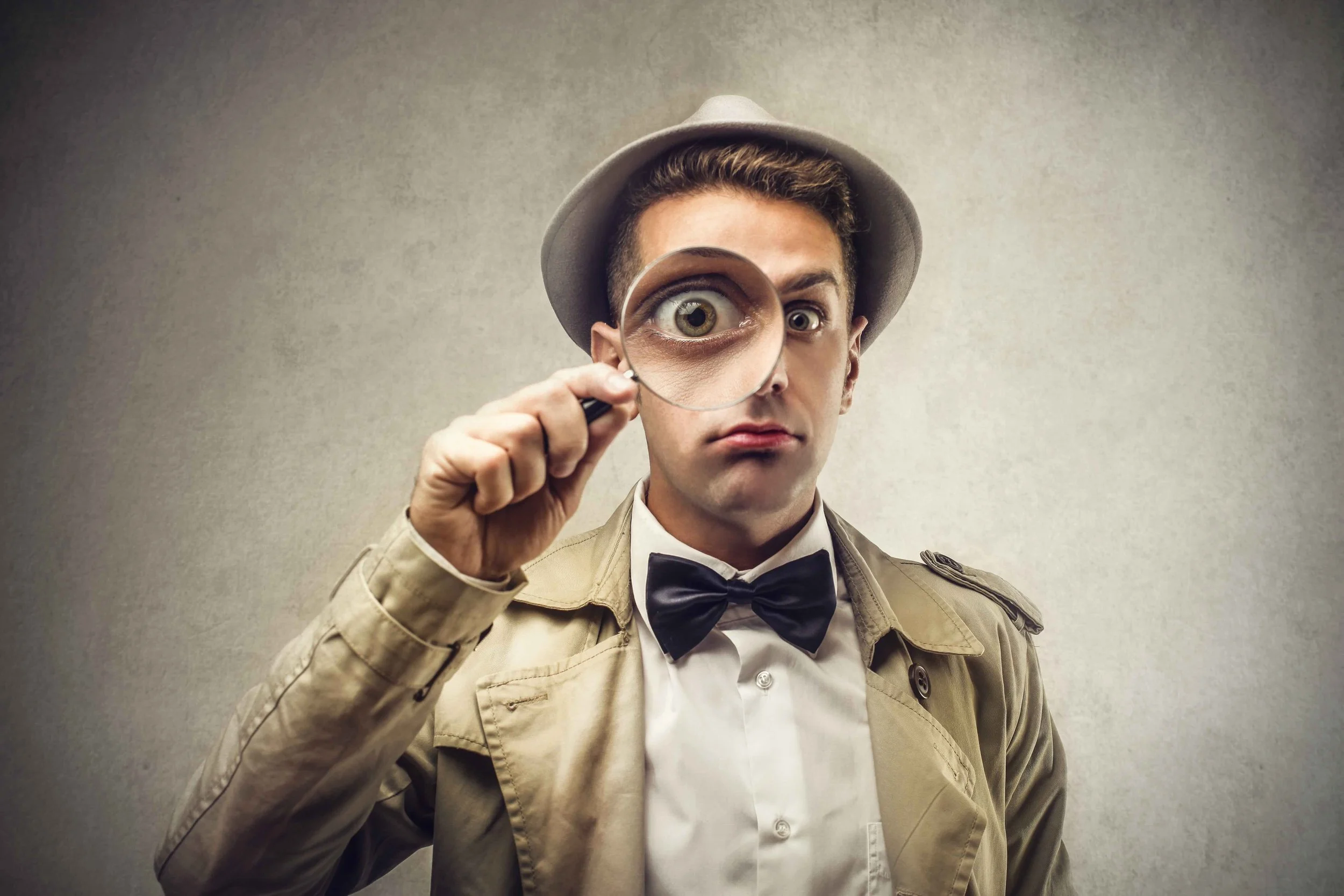 A man in a detective costume holds a magnifying glass in front of one eye, making it appear larger, while wearing a beige trench coat, a white shirt, a black bow tie, and a beige fedora hat, against a plain background.
