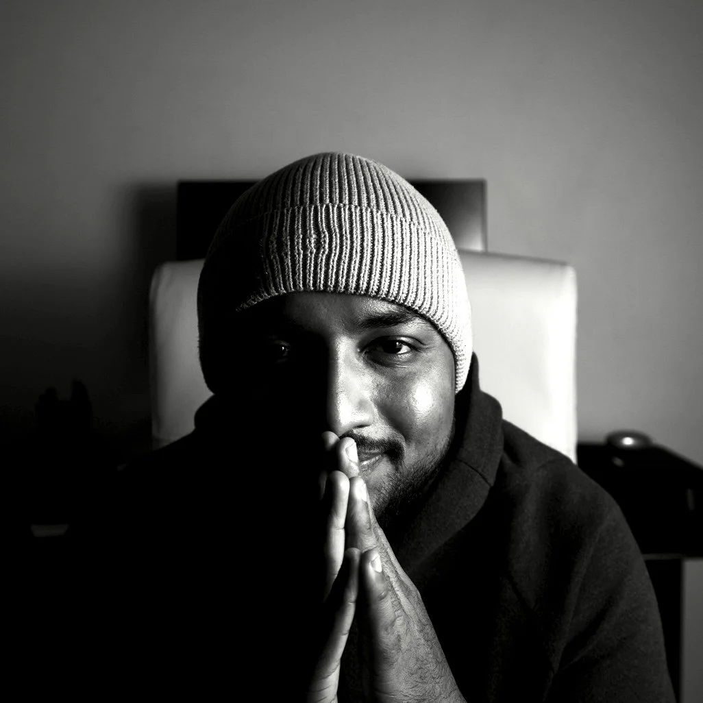 Black and white photo of a young man wearing a knit cap, sitting with hands pressed together near face, in a dimly lit room.