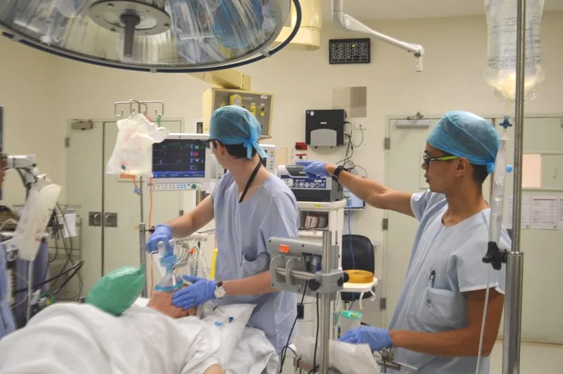 Dr Matthew Ho and another healthcare professional, wearing scrubs, gloves, and surgical caps, are attending to a patient in an operating room with medical equipment and monitors.