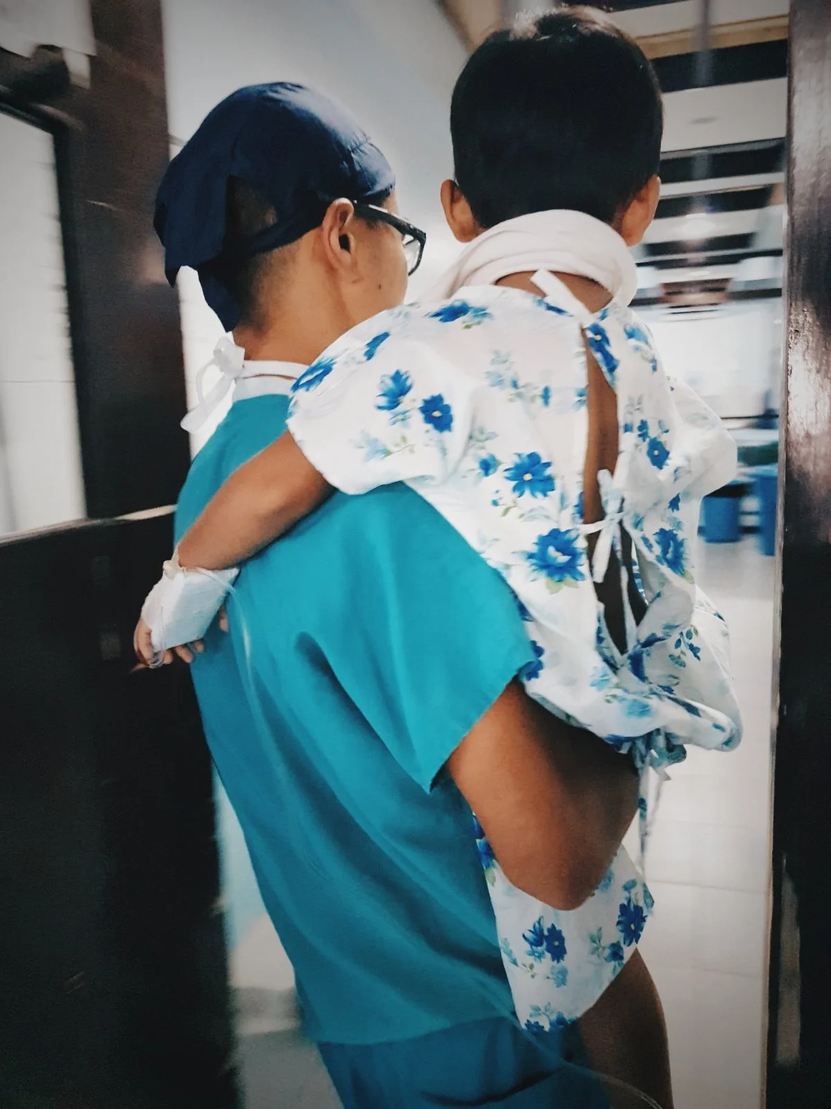 Dr Matthew Ho, in blue medical scrubs and a dark blue surgical cap, carries a young child patient in a hospital hallway.
