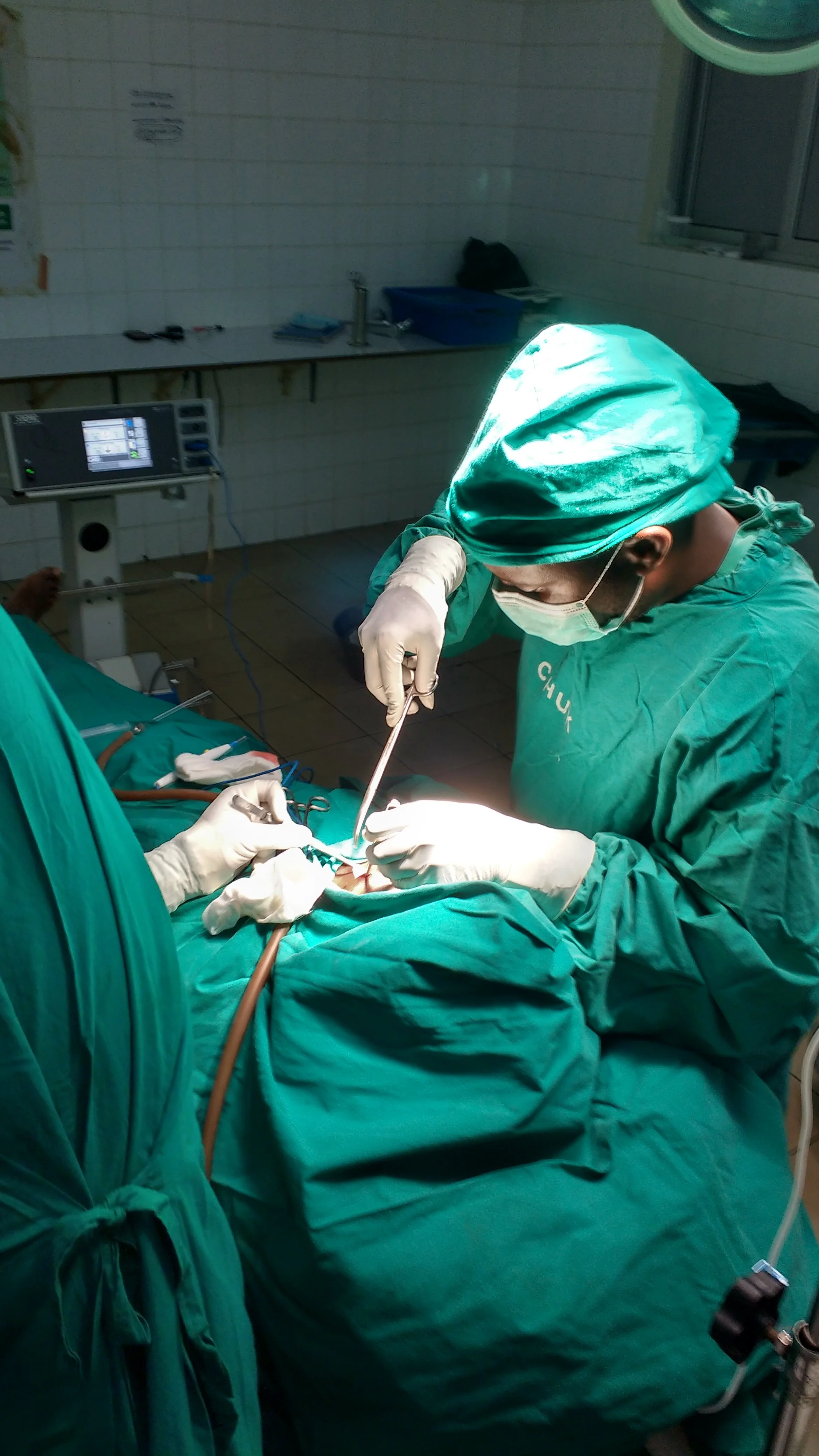 Surgeons performing an operation in an operating room, wearing green surgical scrubs, masks, and gloves, with medical equipment and monitors in the background.
