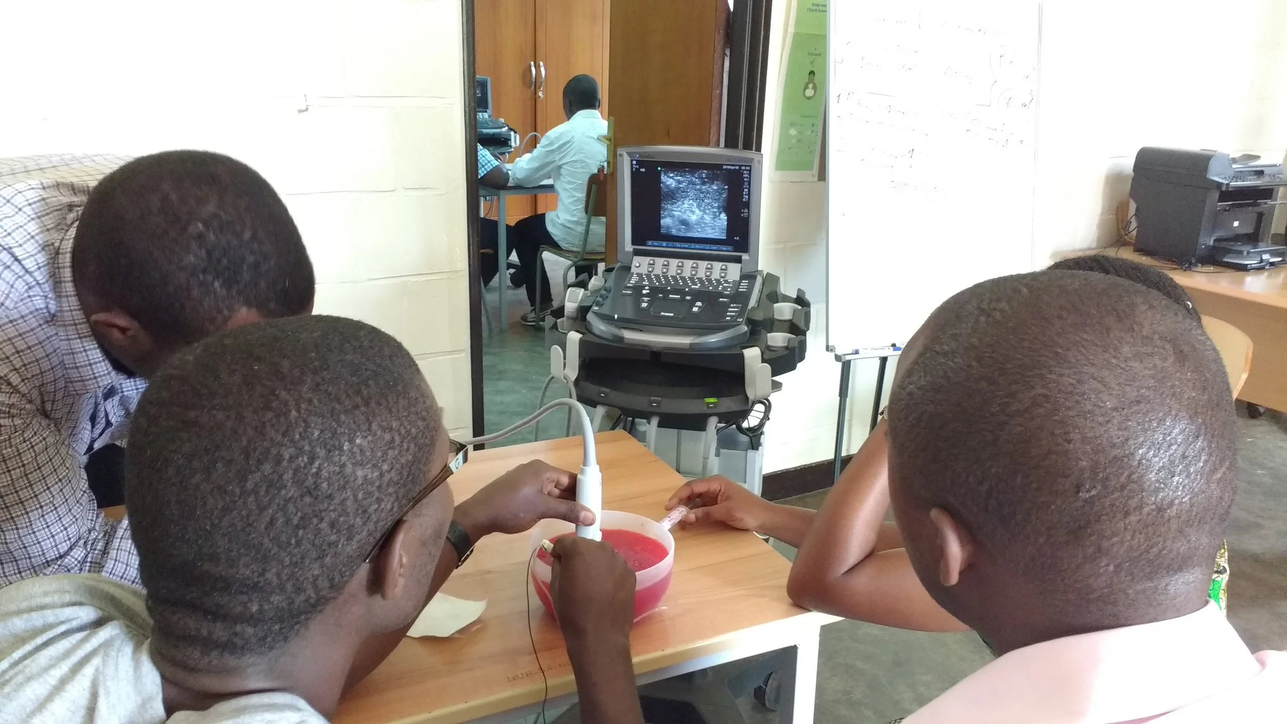 A group of people around a table in a medical examination room, with ultrasound equipment and a medical professional in the background.
