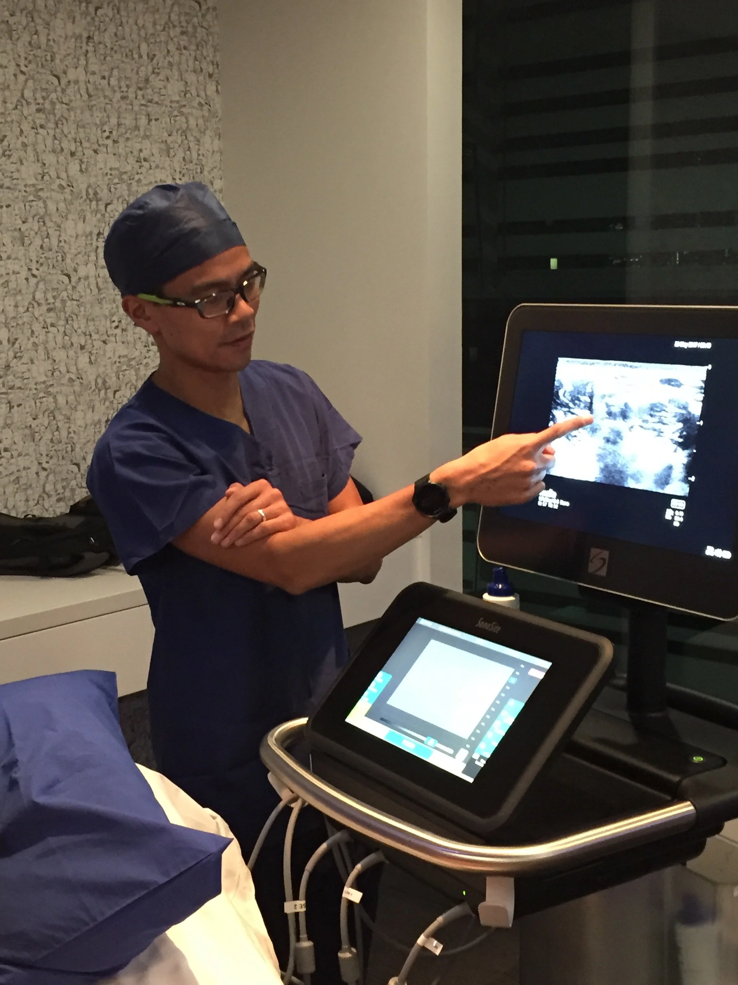 Dr Matthew Ho, dressed in medical scrubs, wearing glasses and a surgical cap, points at a monitor displaying an ultrasound image during a medical procedure in a clinical room.