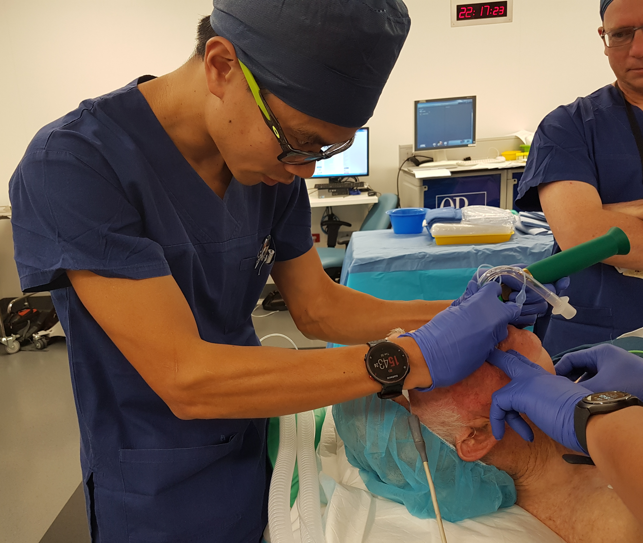 Dr Matthew Ho, anaesthesiologist, wearing scrubs, is intubating a patient lying on an operating table. The patient is covered with a sterile drape. In the background, there are monitors and medical equipment in a clinical setting.