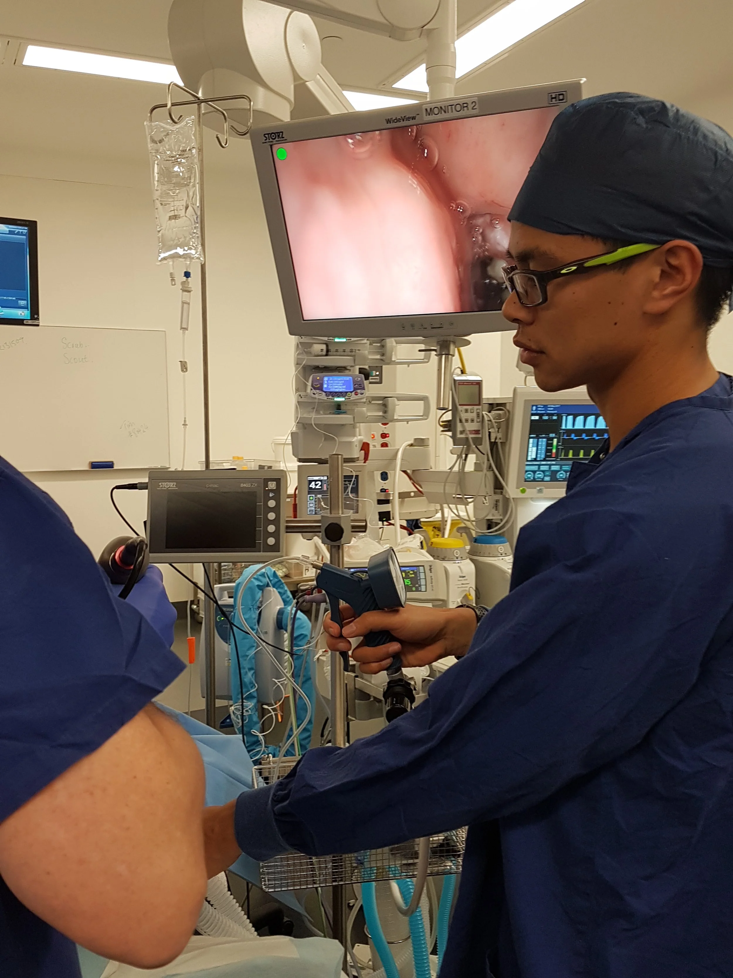 Dr Matthew Ho, anaesthesiologist, supporting a patient in a hospital operating theatre during a medical endoscopy, with monitors displaying internal images of a patient's gastrointestinal tract.