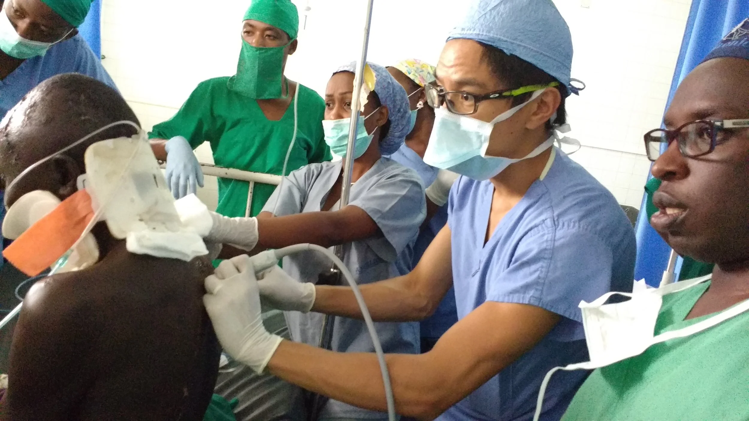 Dr Matthew Ho, specialist anaesthetist, and his medical team performing an ultrasound guided procedure on a patient, with several healthcare professionals in scrubs and masks gathered around.