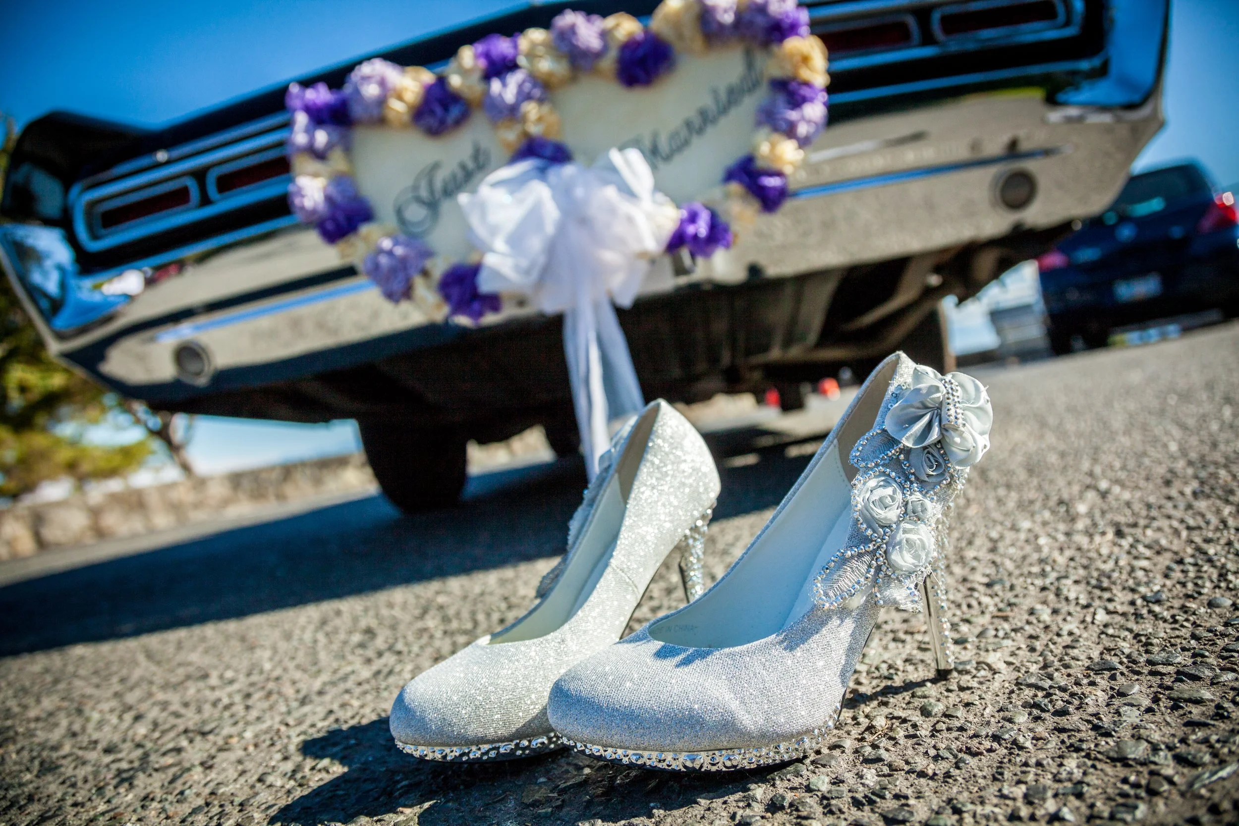White high-heeled shoes decorated with floral embellishments rest on a gravel ground. In the background, a vintage car with a heart-shaped floral wreath and the words 'Just Married' on it is visible.