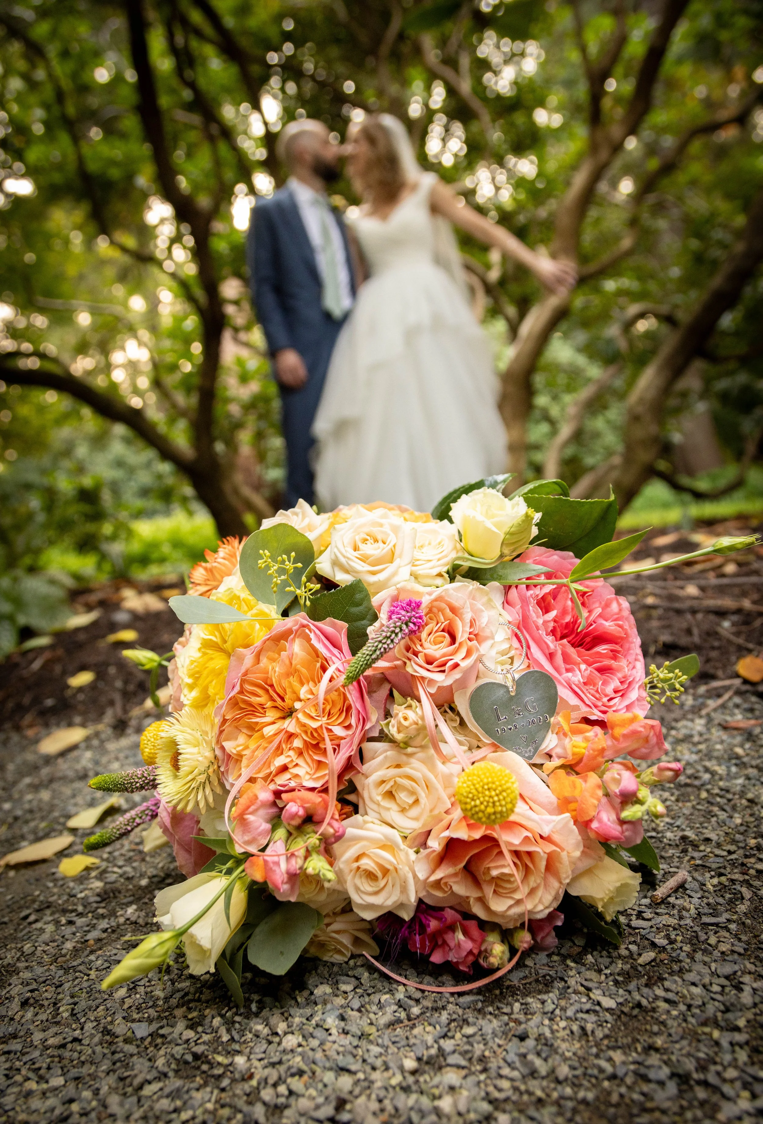 Blurred couple in wedding attire kissing in a wooded outdoor setting, with a bouquet of colorful flowers on the ground in front.