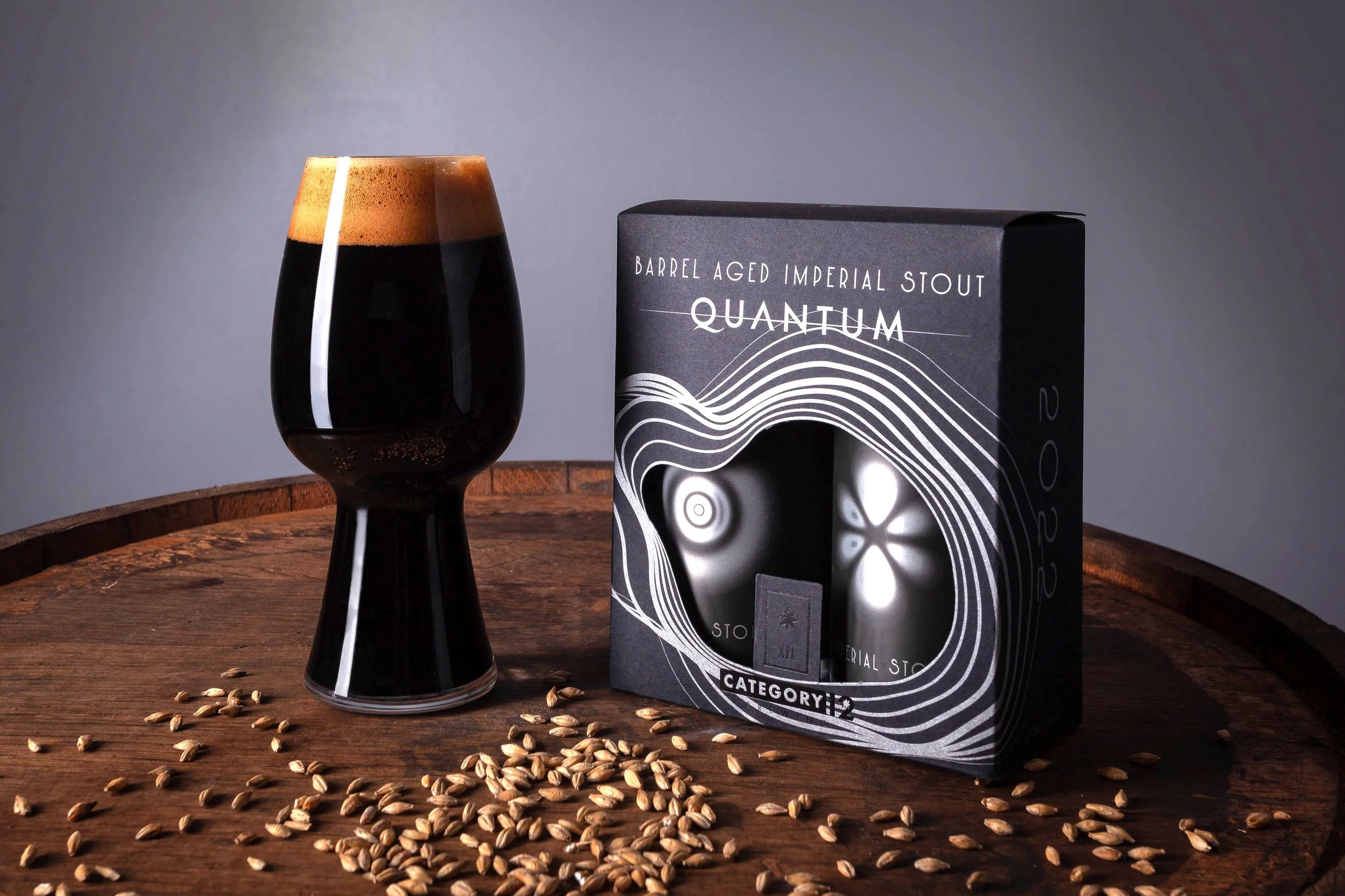 A glass of dark stout beer with a thick tan head next to a black box labeled 'Barrel Aged Imperial Stout Quantum,' with scattered barley grains on a wooden surface.