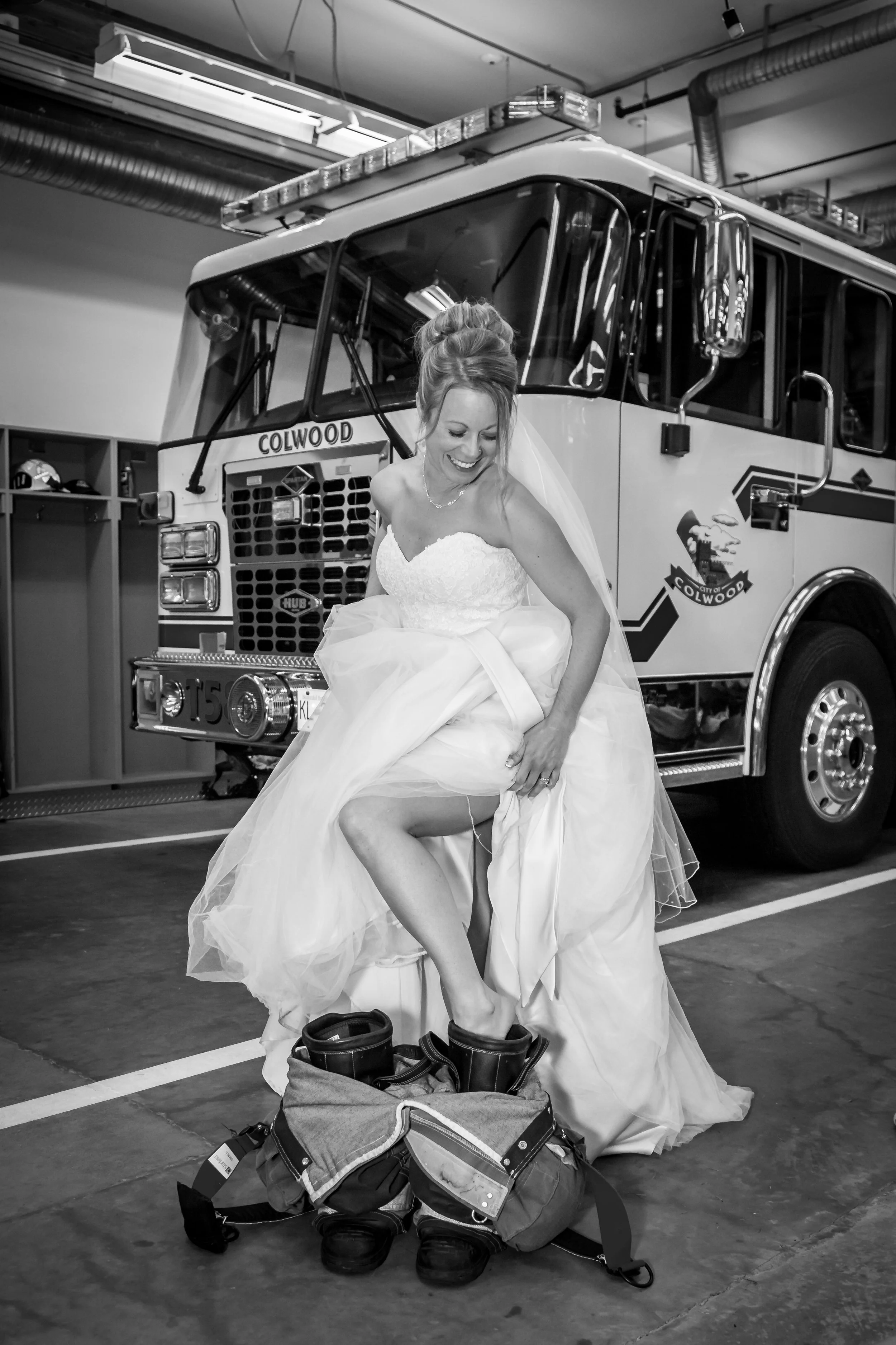 A bride in a wedding dress sitting down in a fire station, smiling and lifting her dress to show her leg, with firefighting gear at her feet and a fire truck in the background.