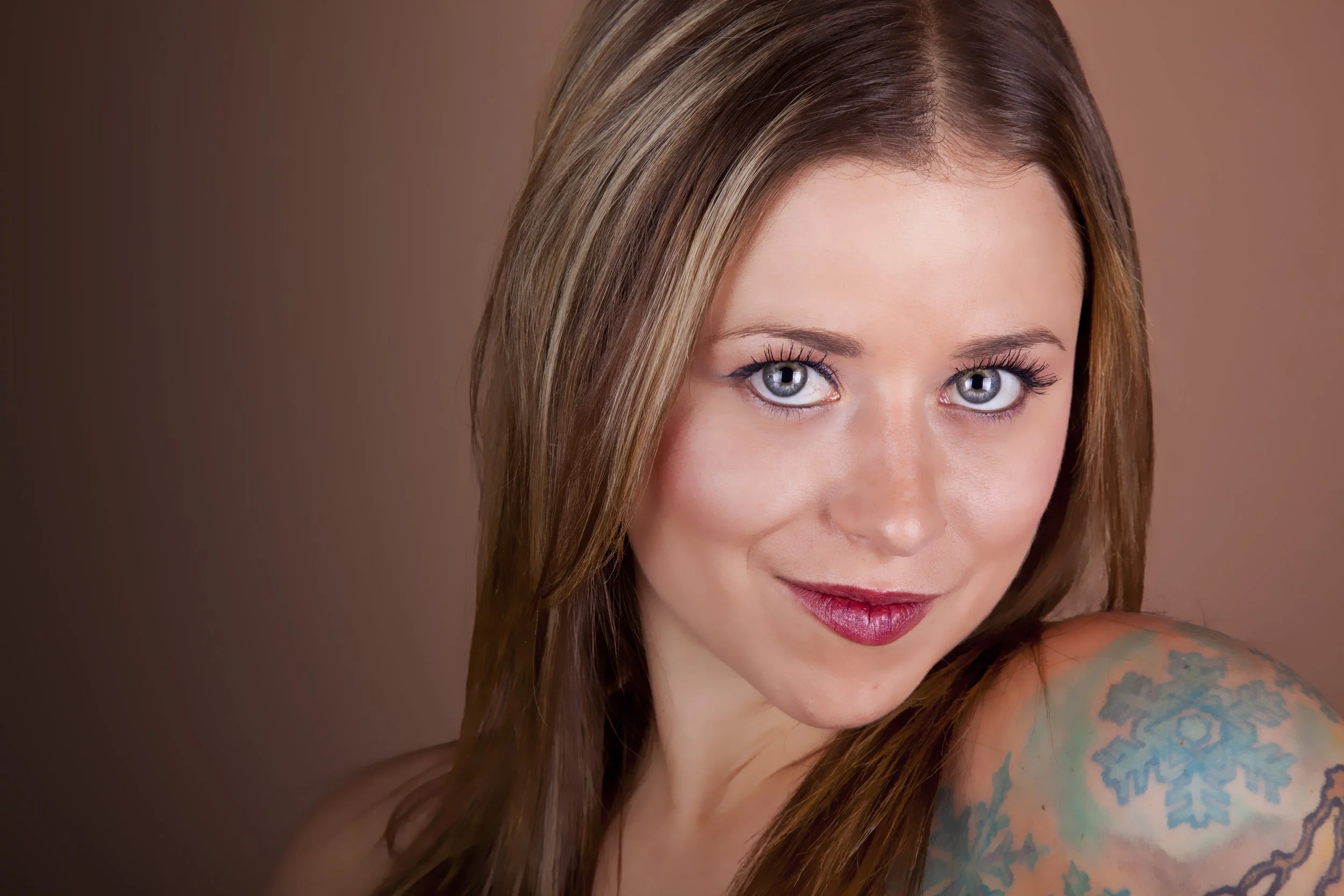 Close-up of a woman with red hair, blue eyes, wearing makeup, smiling, with bare shoulders and a colorful tattoo on her right shoulder.