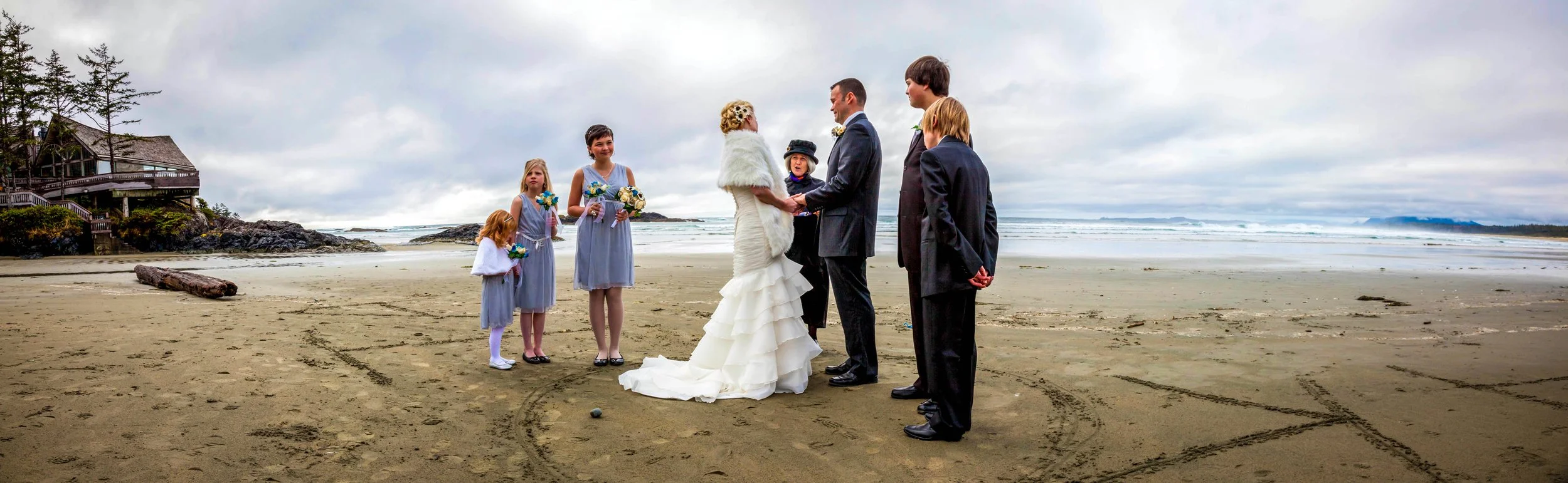 A beach wedding ceremony with a bride and groom standing face to face, holding hands, surrounded by two bridesmaids and two groomsmen, under a cloudy sky near the ocean.