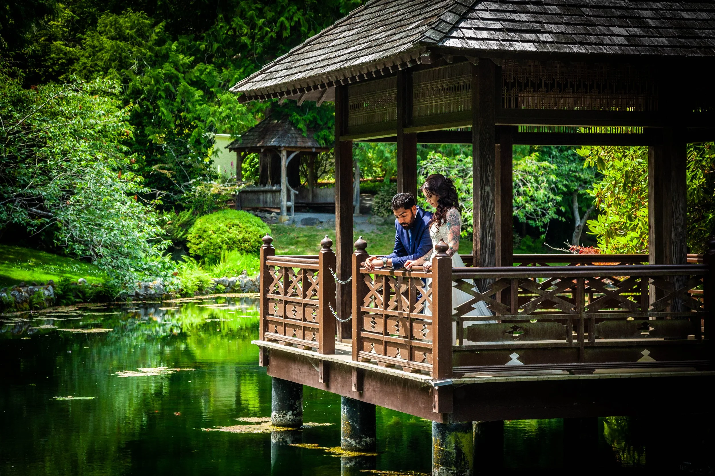 A couple in wedding attire standing on a wooden balcony over a pond, surrounded by lush greenery and trees.