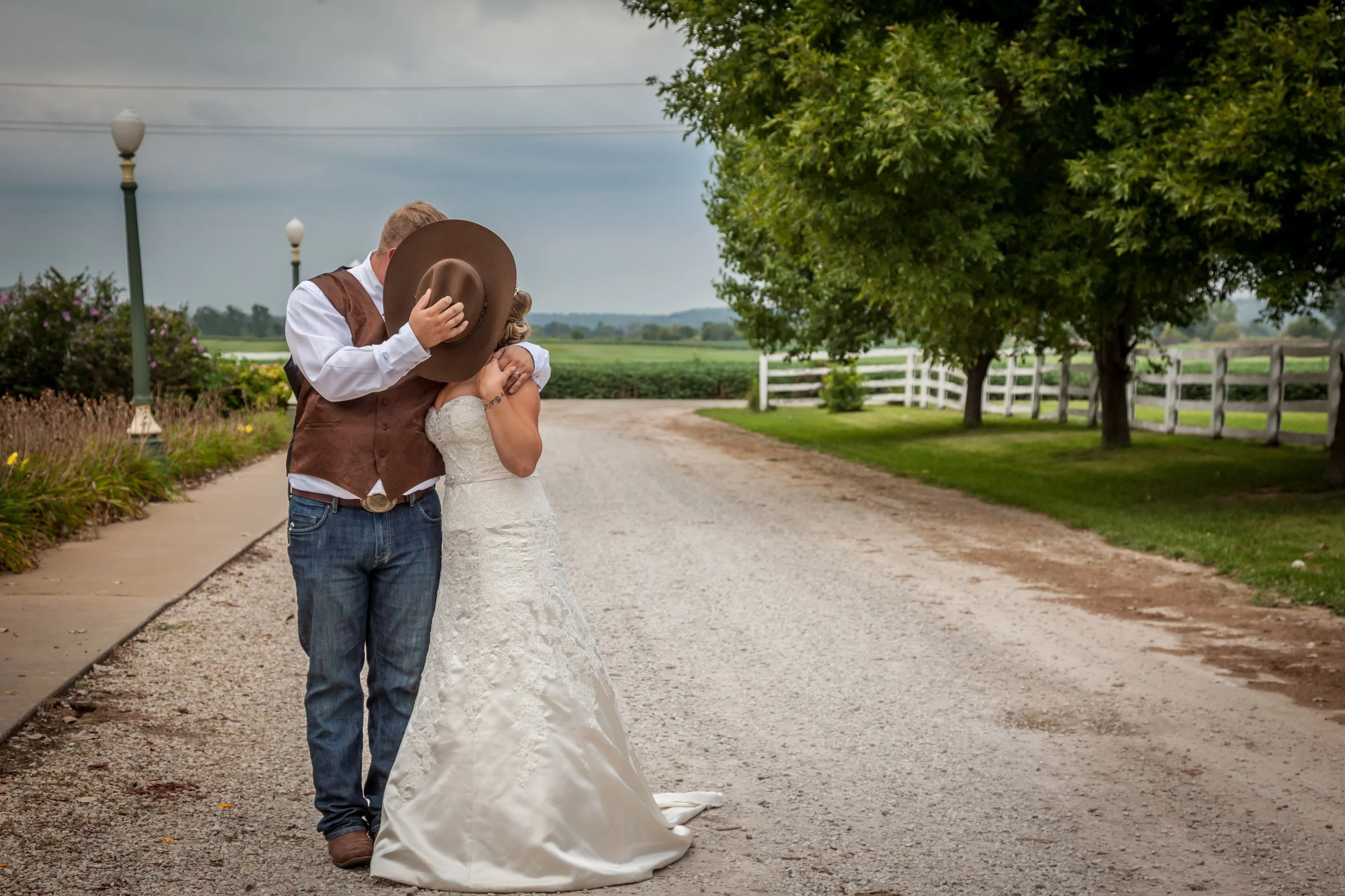 A couple on their wedding day sharing a kiss outdoors on a gravel path, with trees, a white fence, and cloudy sky in the background.