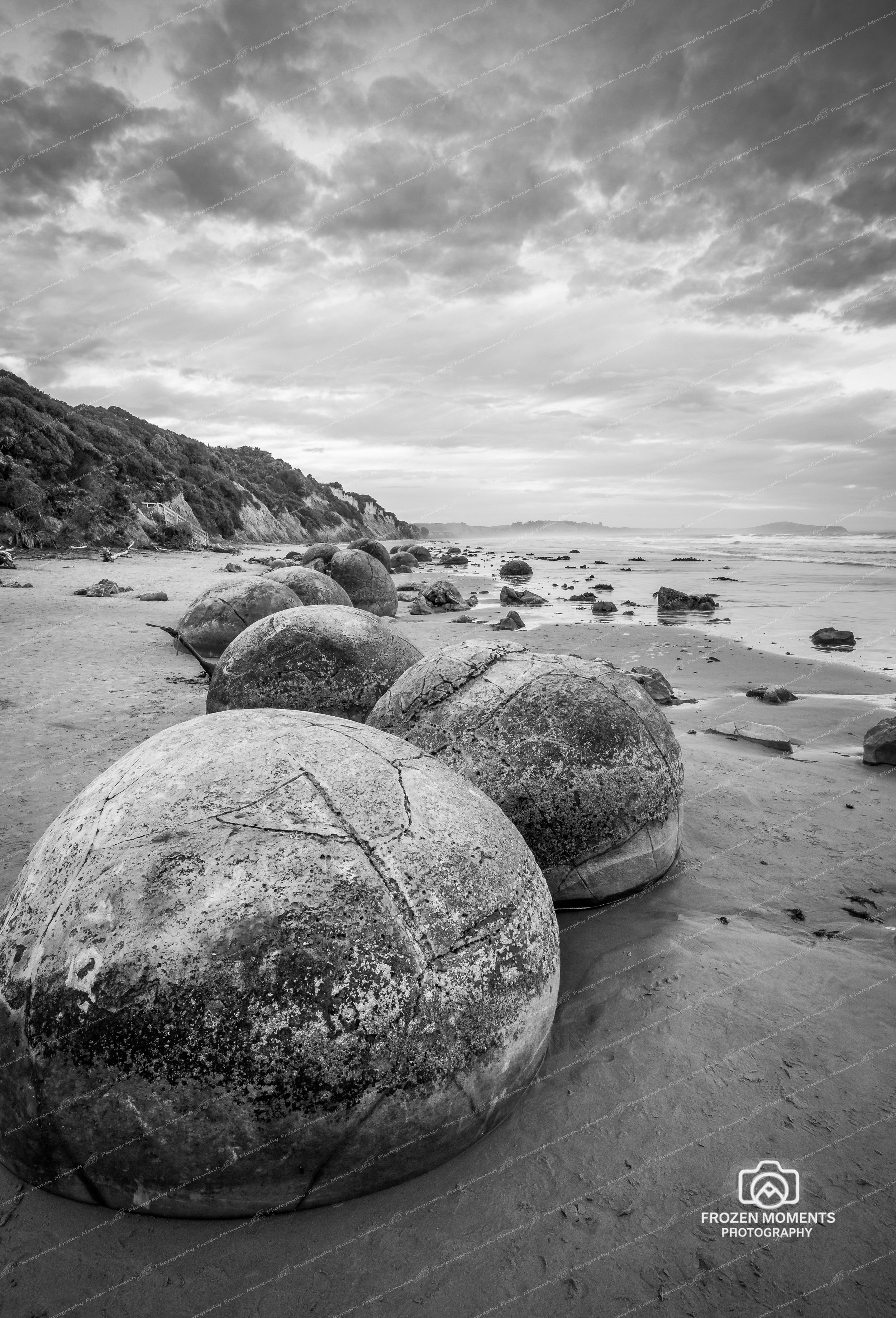 Black and white photo of large rounded rocks on a sandy beach, with smaller rocks scattered along the shoreline. Hills covered with dense trees are in the background, and the sky is filled with clouds.
