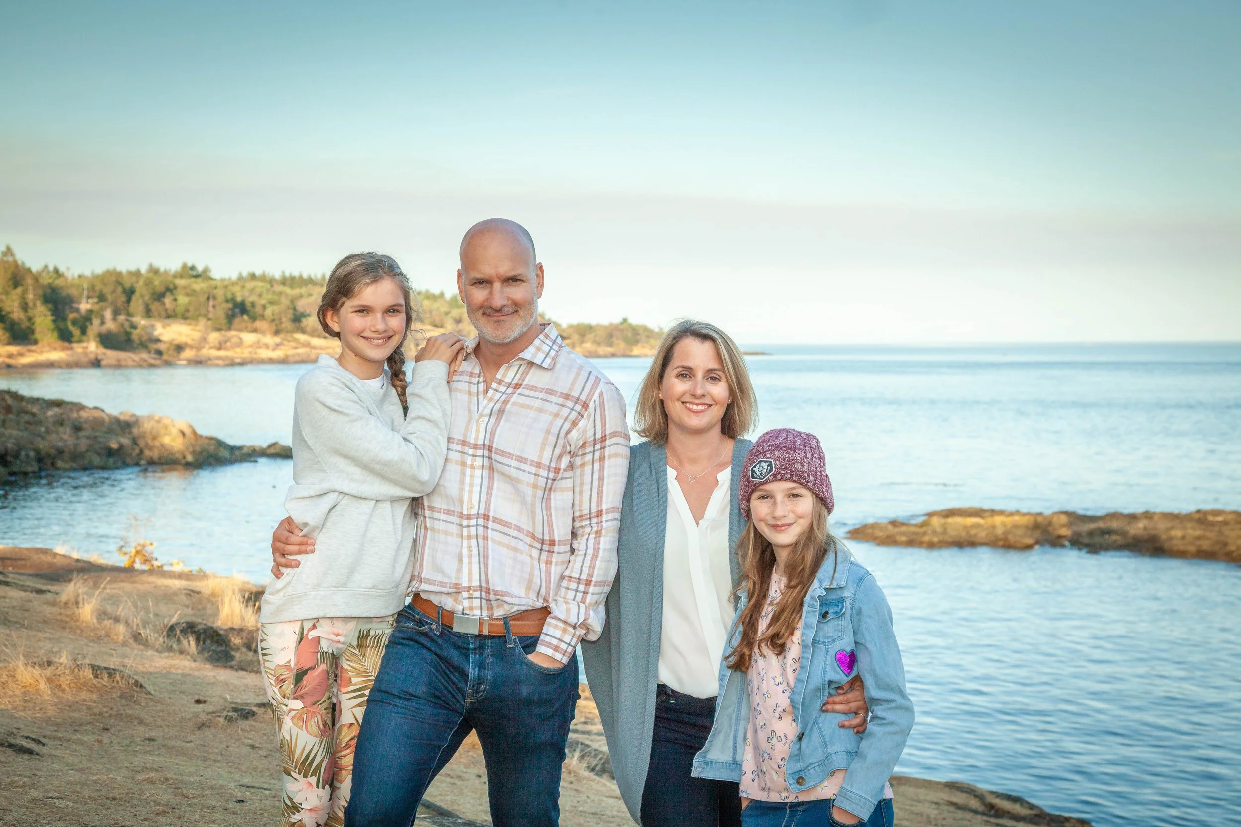 A family of five standing on a rocky shoreline near the water, with a forest and blue sky in the background.