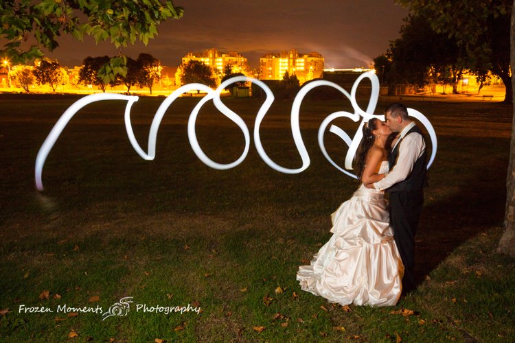 A bride and groom kissing outdoors at night, with a light-painted heart and the word 'love' in the background, illuminated by long-exposure photography.