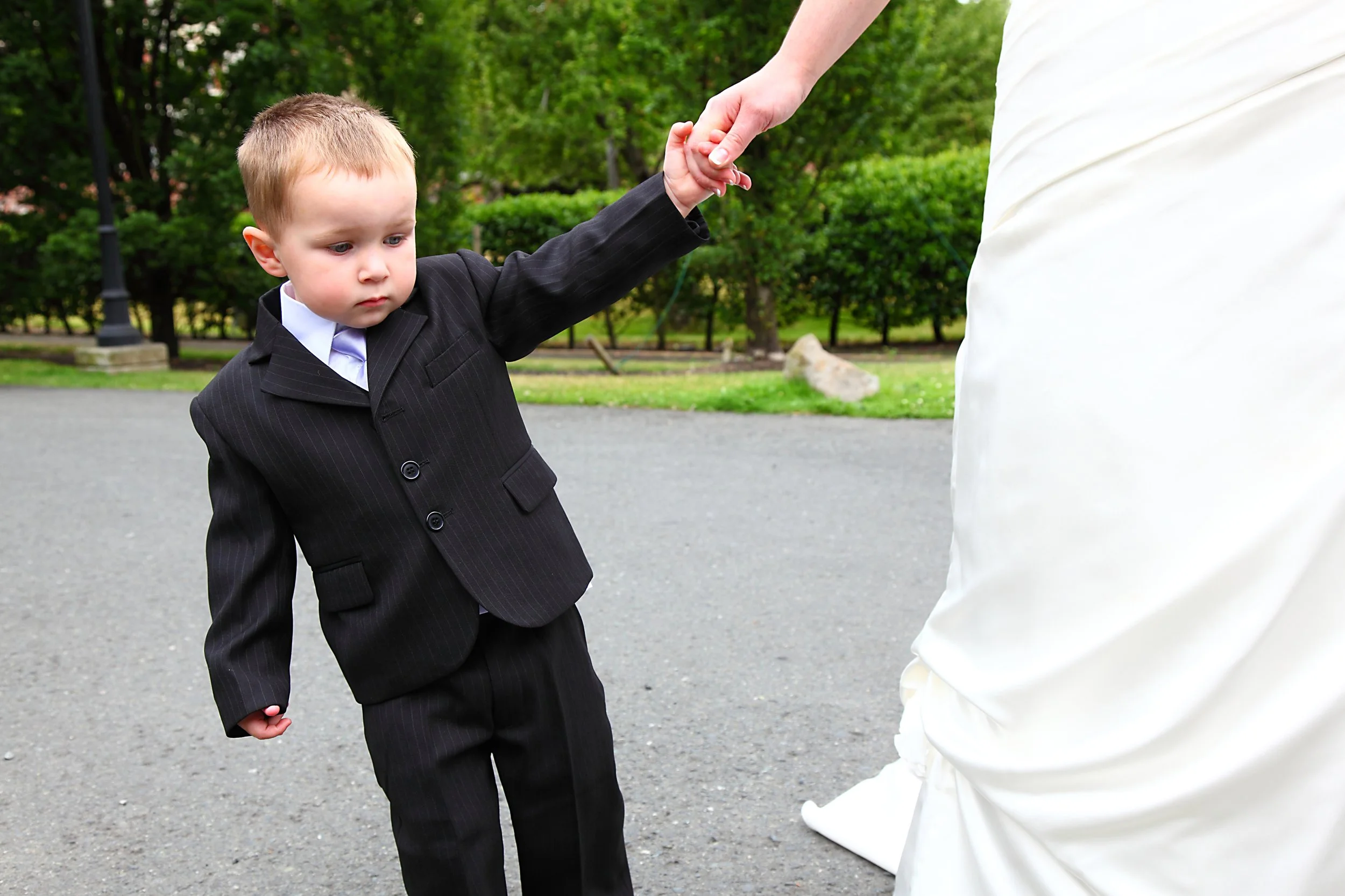 A young boy in a suit holding hands with a person in a white dress, outside in a park or garden area.