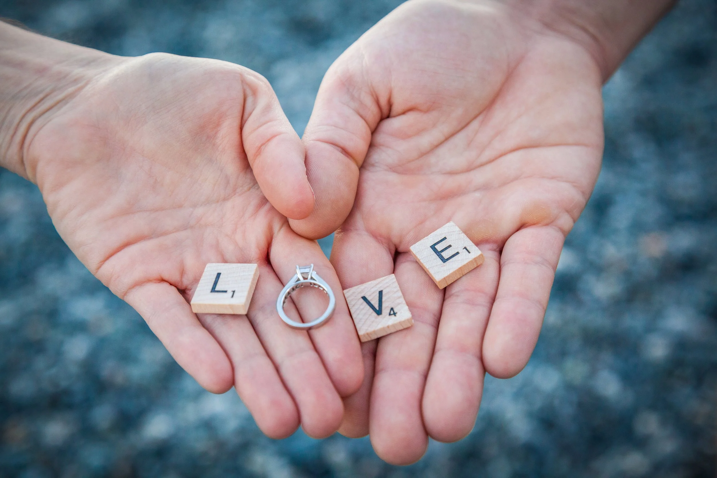 Close-up of hands holding Scrabble tiles spelling out 'LOVE' and a silver ring.