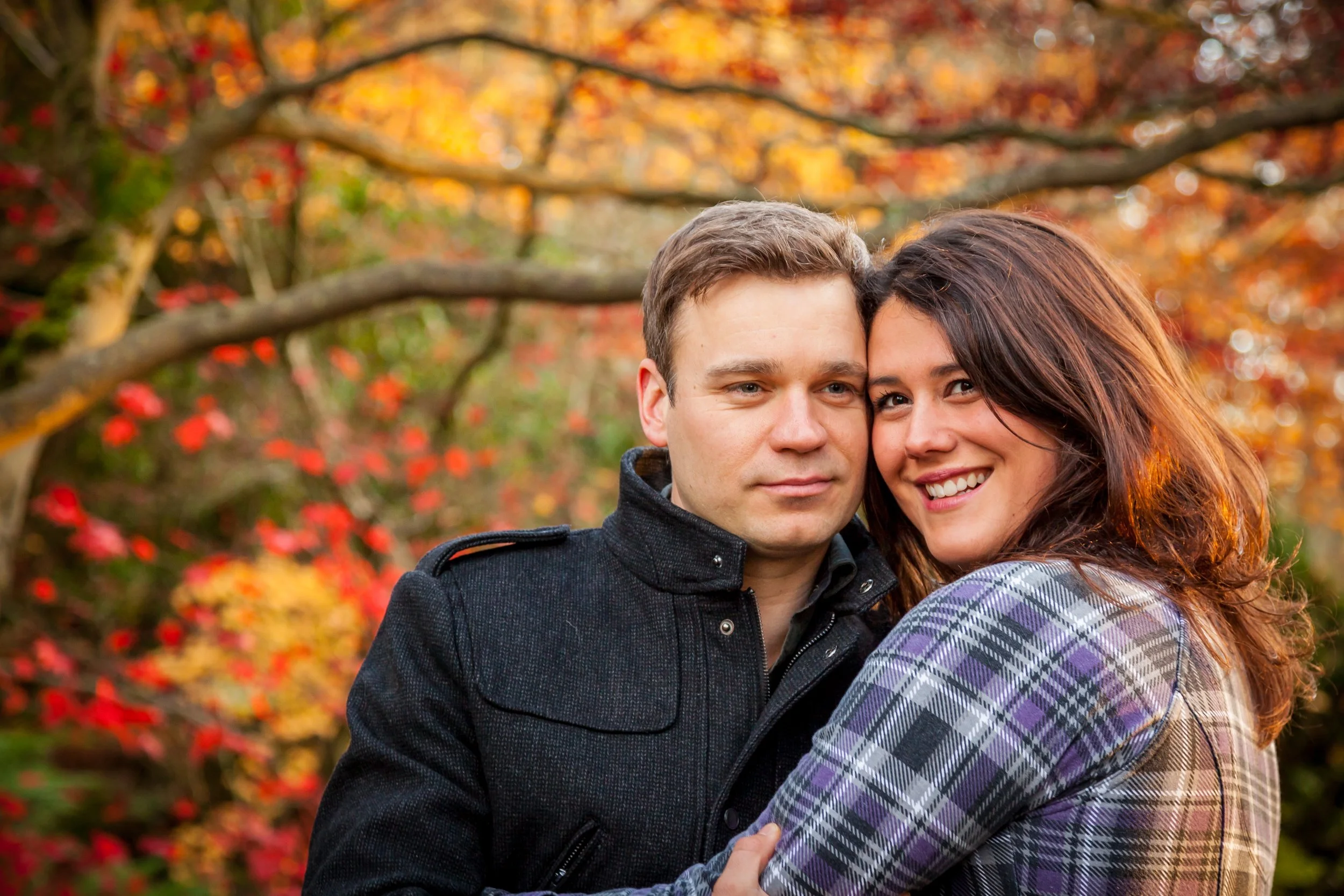 A young couple embracing outdoors in a park during autumn with colorful fall foliage in the background.