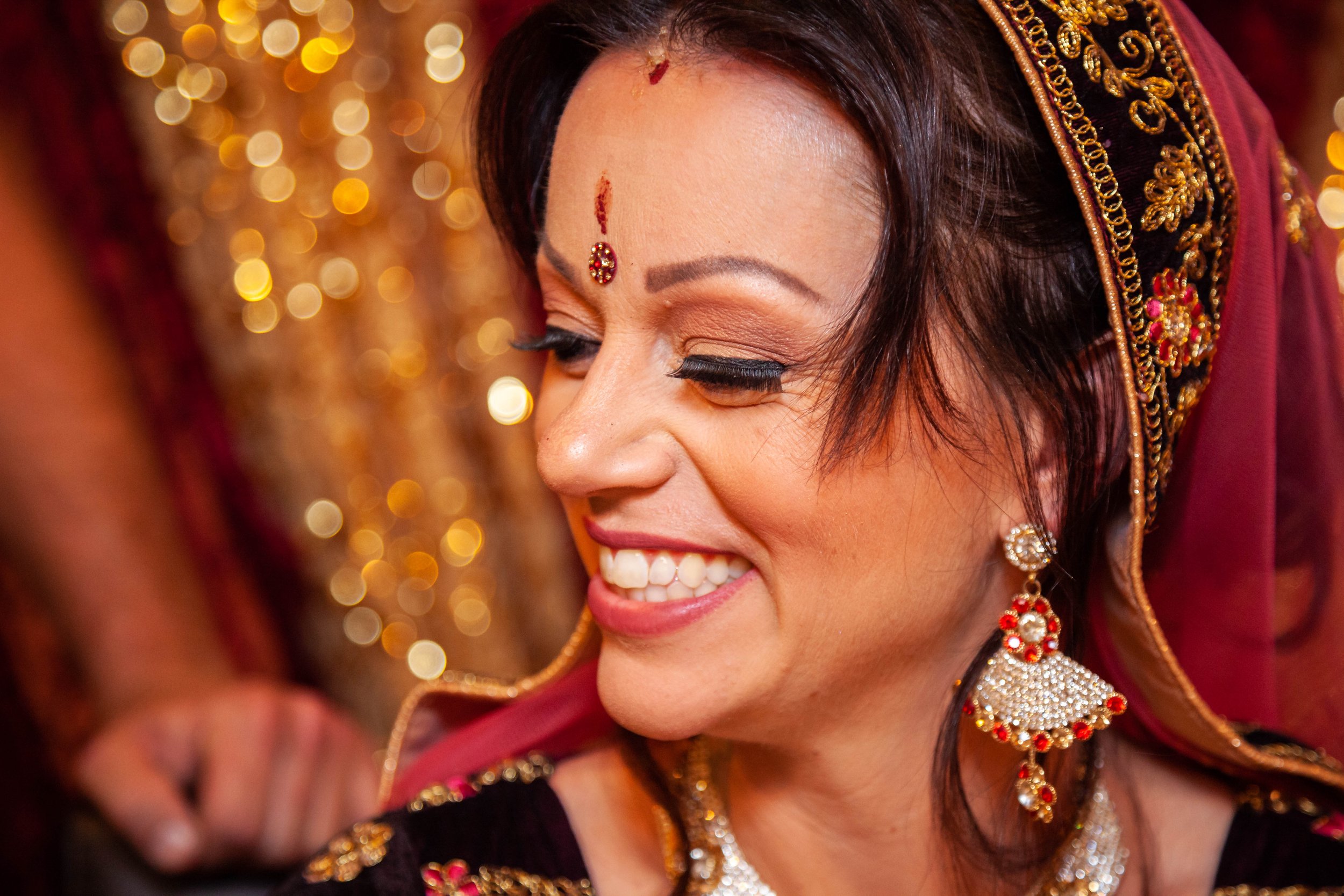 Close-up of a smiling woman dressed in traditional Indian attire with jewelry, makeup, and decorative accessories, with a background of bokeh lights.