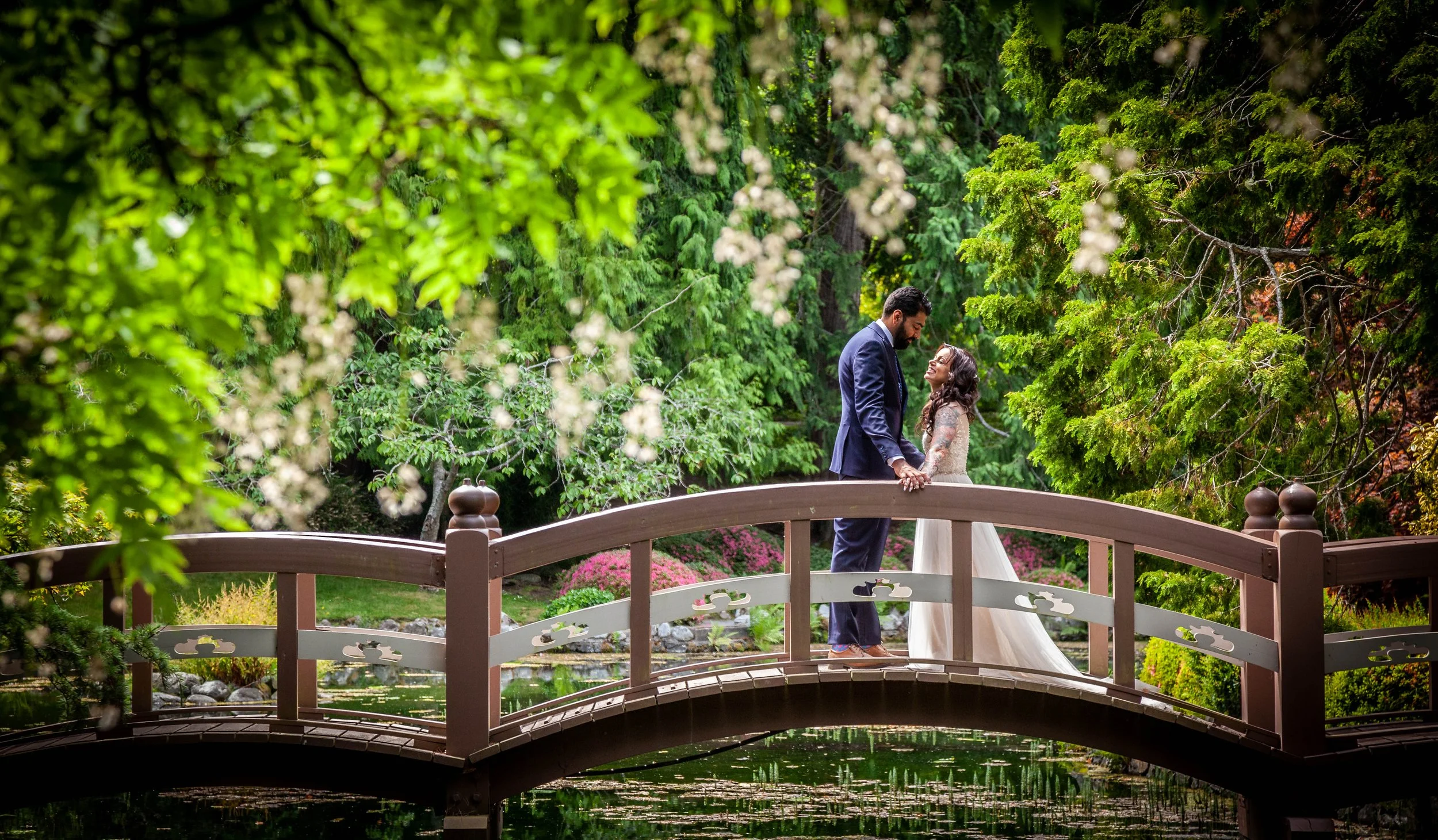 A couple in wedding attire holding hands and smiling on a small wooden bridge in a lush green garden.