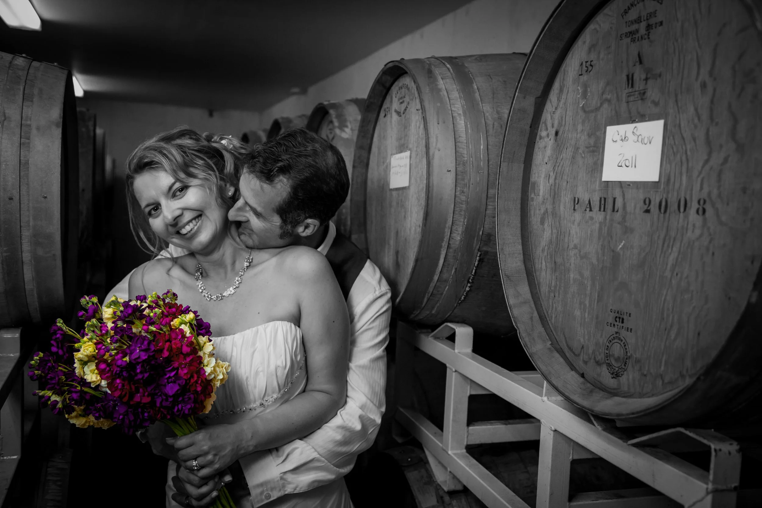 A smiling woman in a wedding dress holding a colorful bouquet of flowers, embracing a man in a tuxedo, standing in a wine cellar with large barrels in the background.