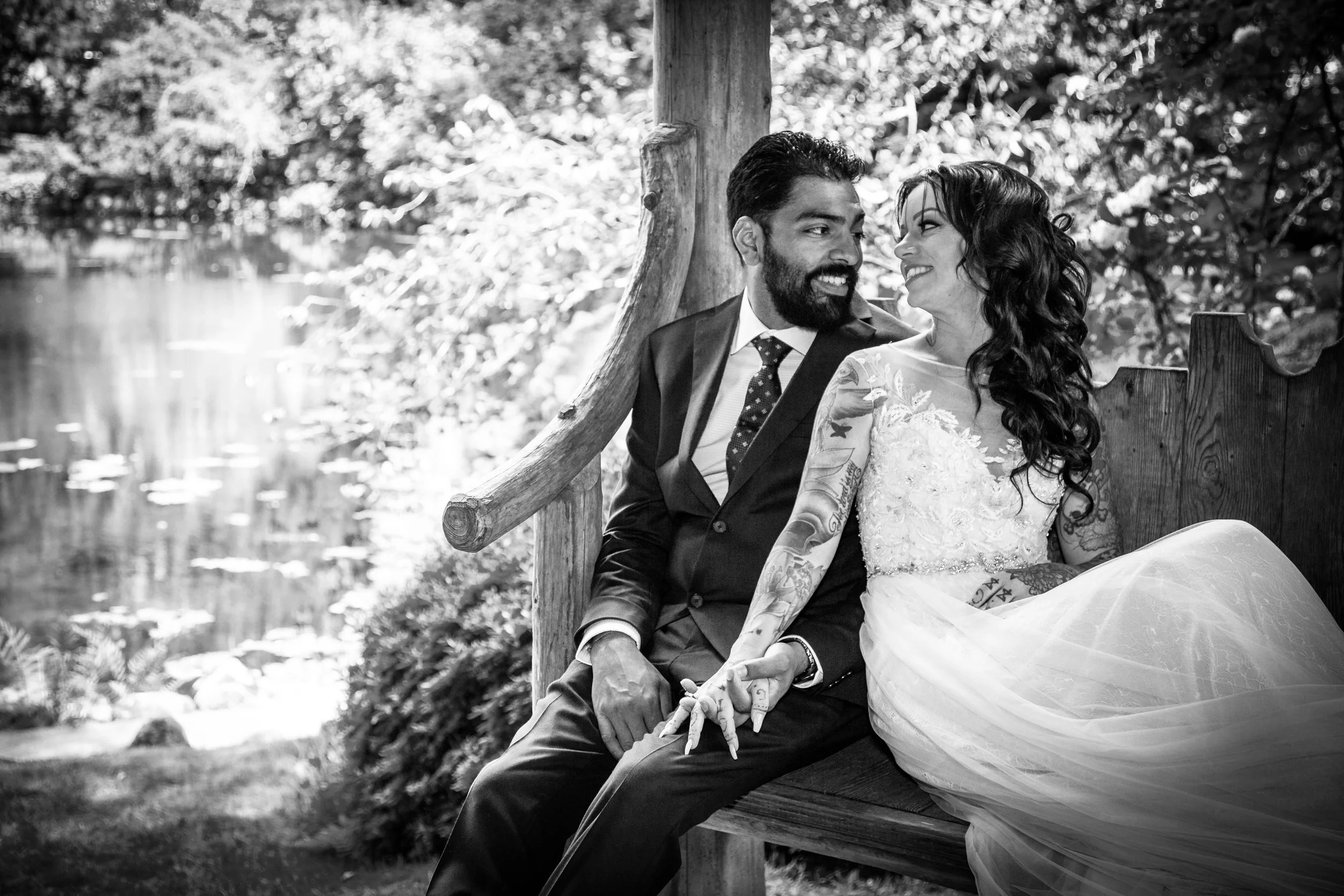 A black-and-white photo of a couple sitting on a wooden bench outdoors, smiling at each other. The man has a beard and short hair, wearing a suit, and the woman has long curly hair, wearing a wedding dress. They are holding hands, with a body of wate