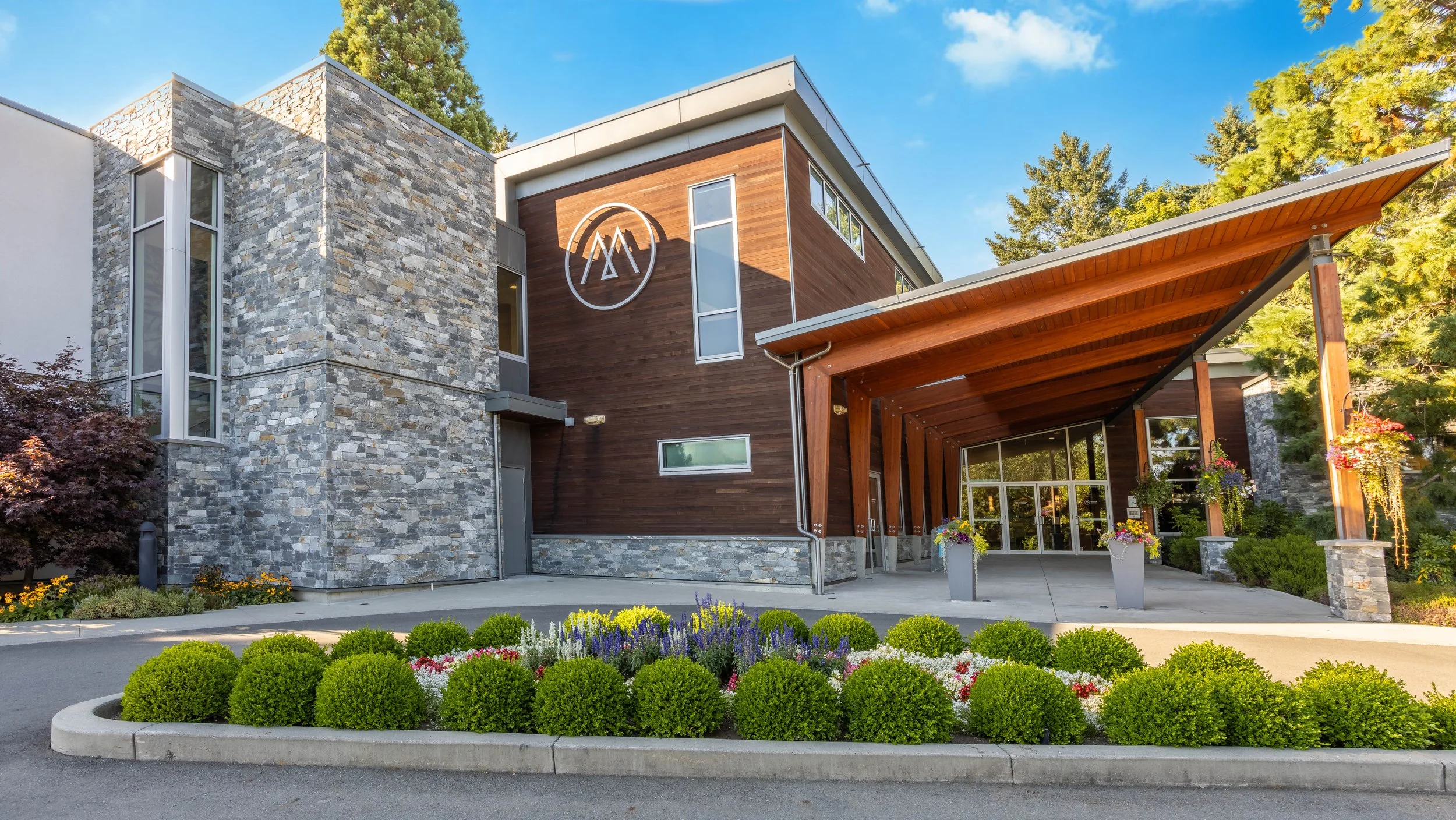 Modern building with stone and wood exterior, surrounded by colorful flower garden and trees, under blue sky.