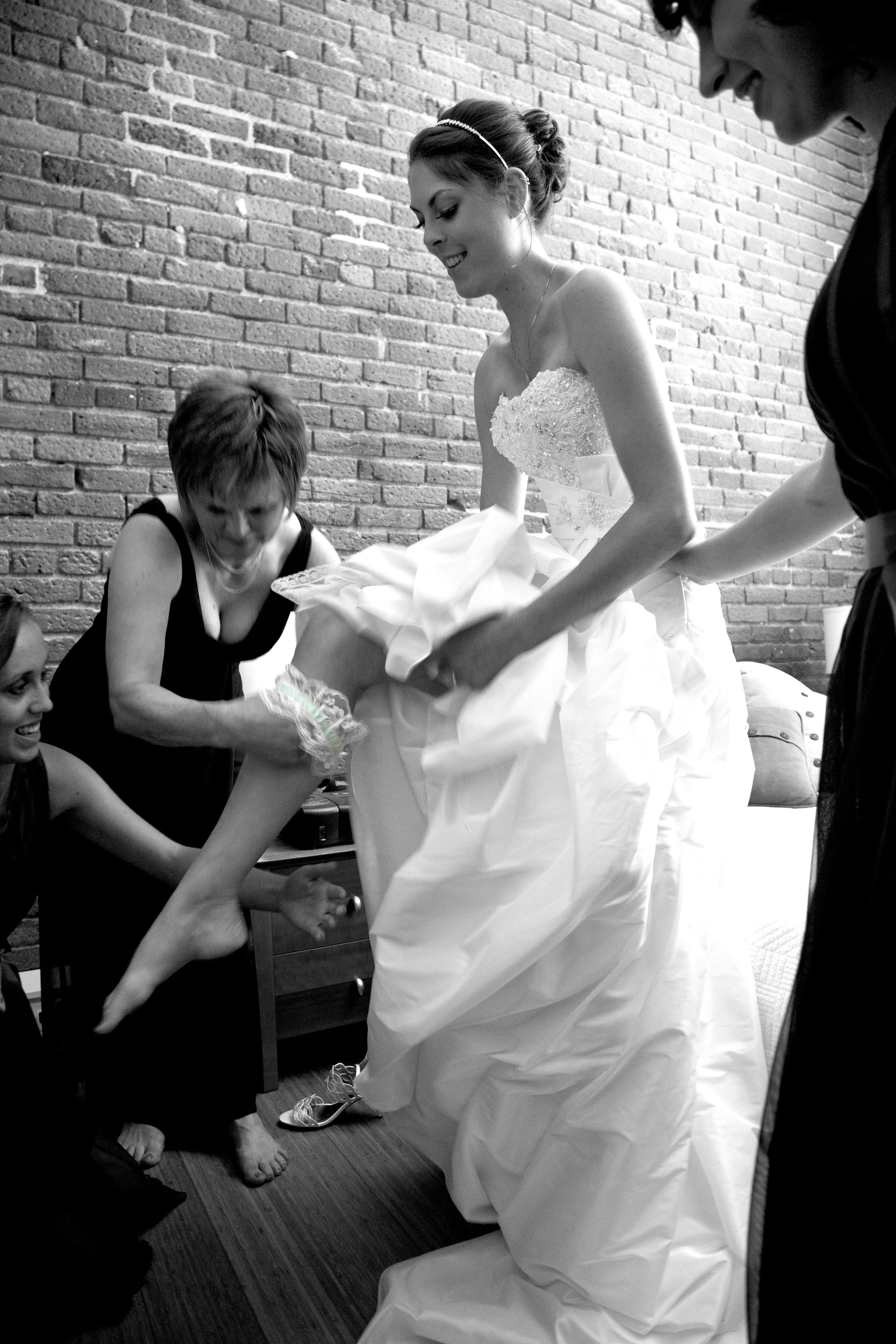 Black and white photo of a bride in a wedding dress, being assisted by a woman and a man, while others look on. They are in a room with a brick wall.