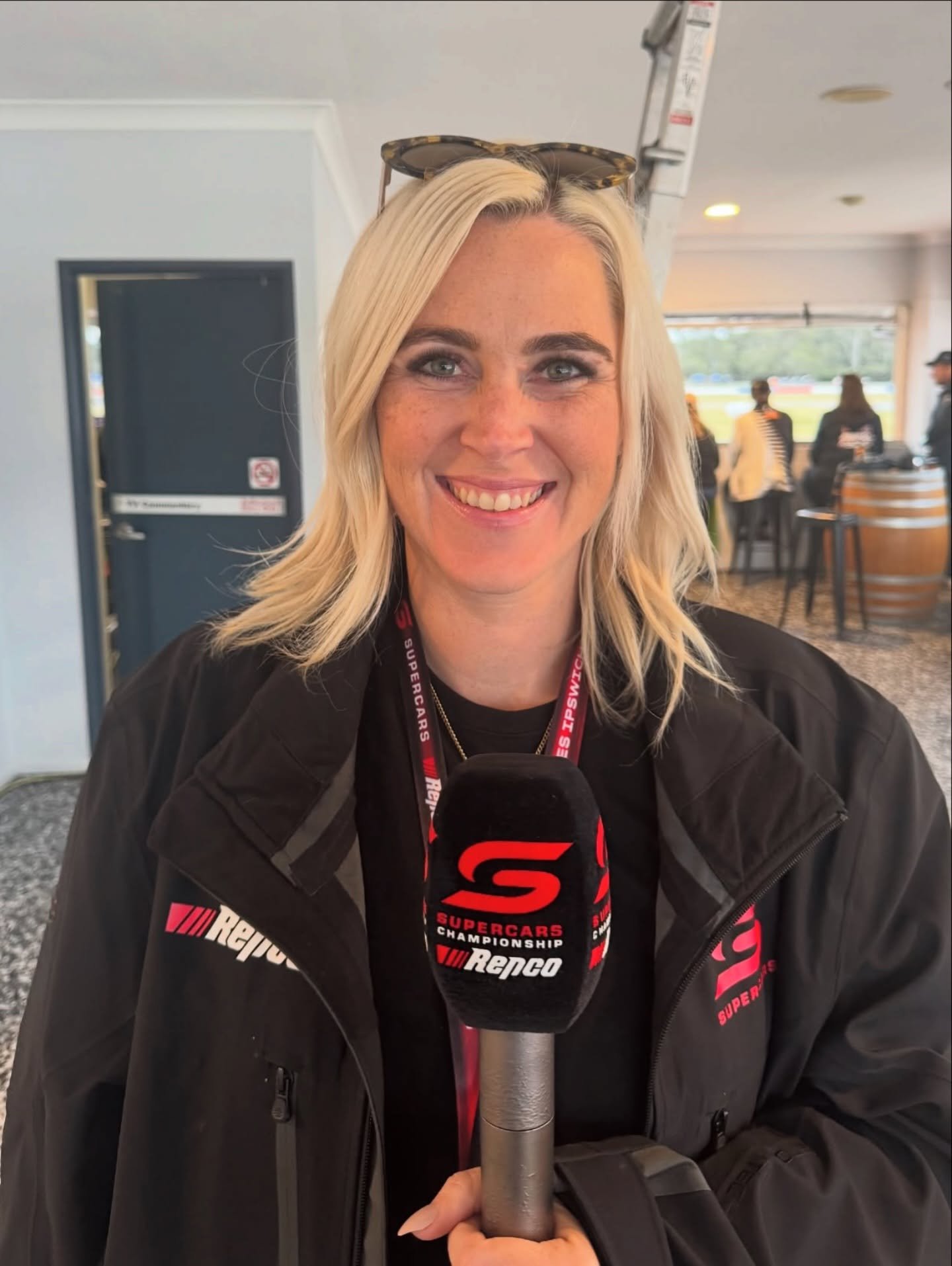 Smiling woman with blonde hair holding a microphone, wearing a black jacket with racing logos, inside a building with large windows showing a racetrack outside.