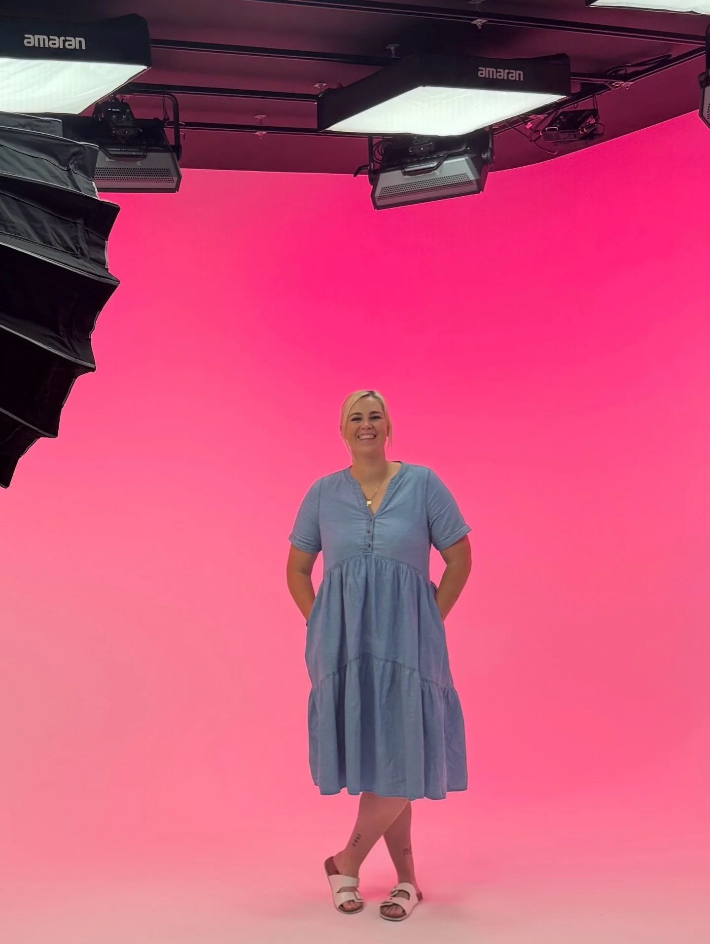 A woman in a blue dress standing on a pink background during a photoshoot, with studio equipment visible above and to the side.