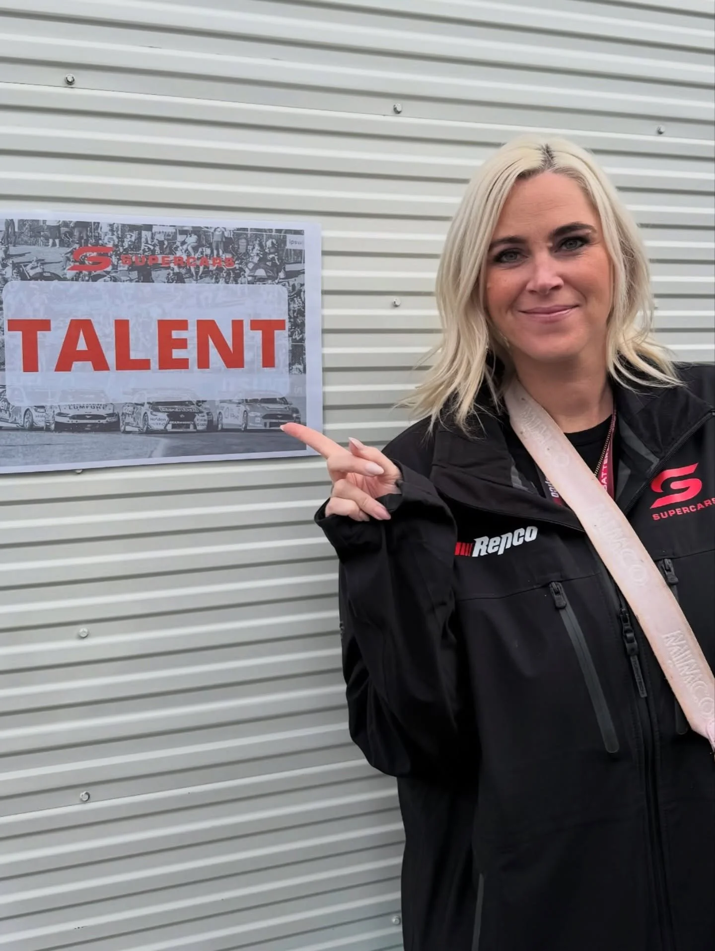 A woman with blonde hair wearing a black jacket pointing at a sign that says "TALENT" with a background of cars and a metal wall.