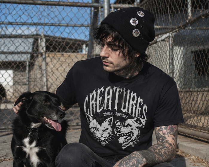 A young man with tattoos and dark hair, wearing a black beanie with pins and a black T-shirt with a graphic design and the word 'CREATURES,' sitting next to a black dog with white markings, in an outdoor fenced area.