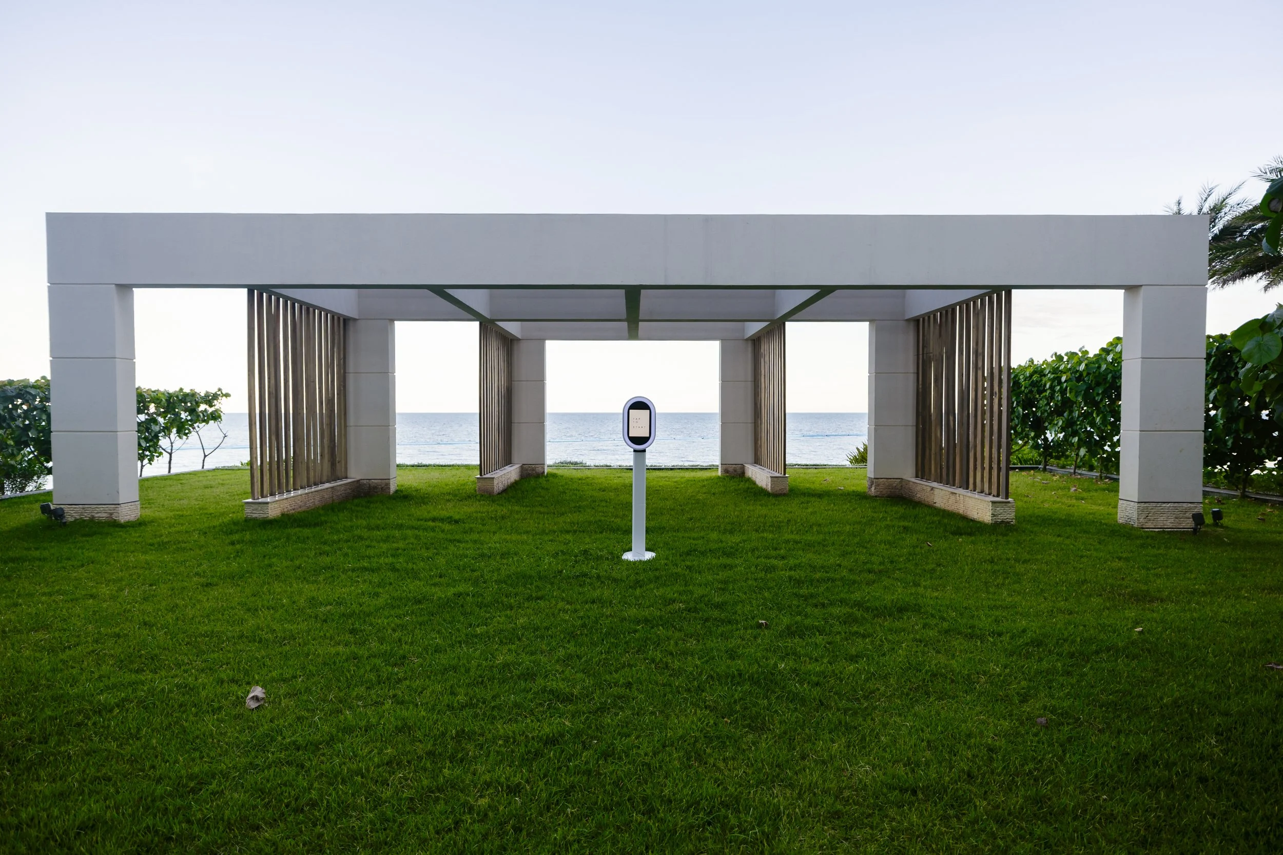 A modern outdoor pergola structure with white and wooden panels, situated on a grassy area facing the ocean, with a small white sign in front with the 1881 studio photo booth.