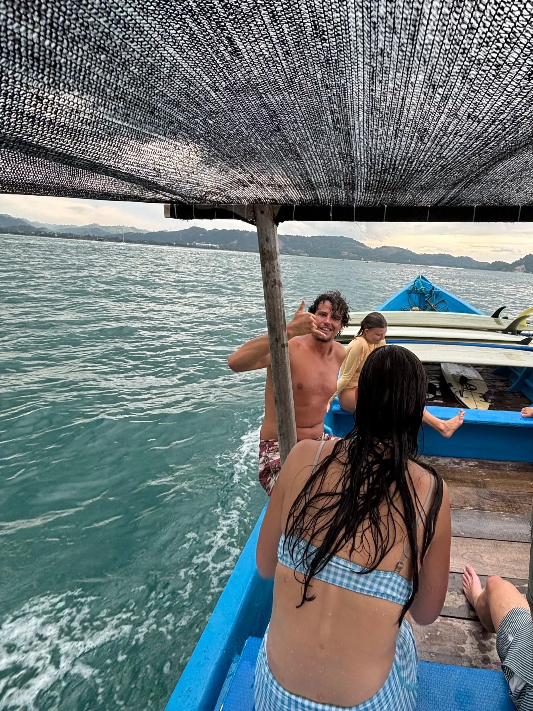 People on a boat in the water, with a man making a hand gesture, some individuals sitting, and mountains in the background.