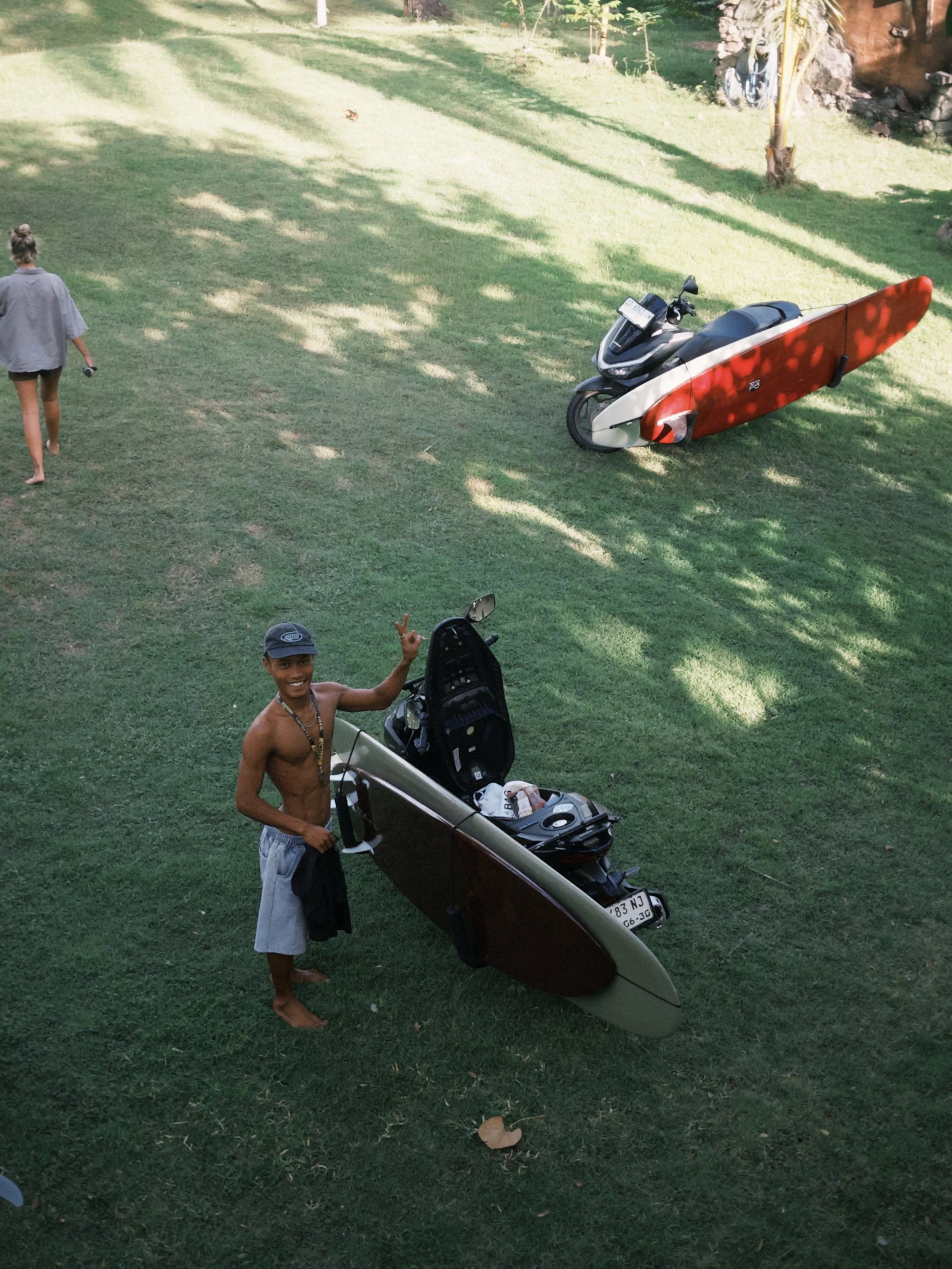 A young shirtless man wearing shorts and a cap is standing on the grass, smiling and waving at the camera. Behind him is a small boat with an open storage compartment. A woman with her hair tied up, wearing a loose shirt and shorts, is walking away, holding a small item in her hand. In the background, a black scooter with a red and white surfboard attached to it is parked on the grass. There are a few trees, rocks, and bicycles in the far background under bright sunlight.